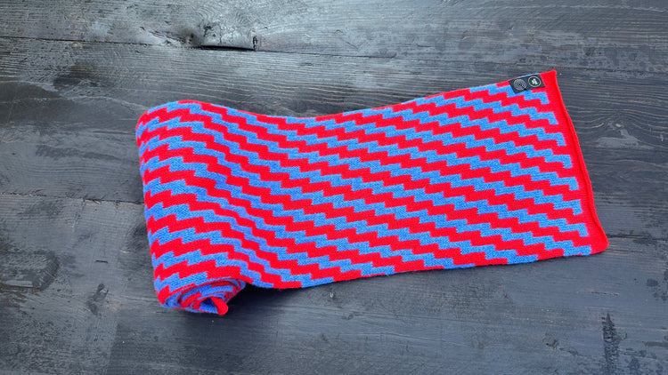 Red and blue rick rack zigzag pattern merino lambswool scarf, partially unrolled on dark wood.