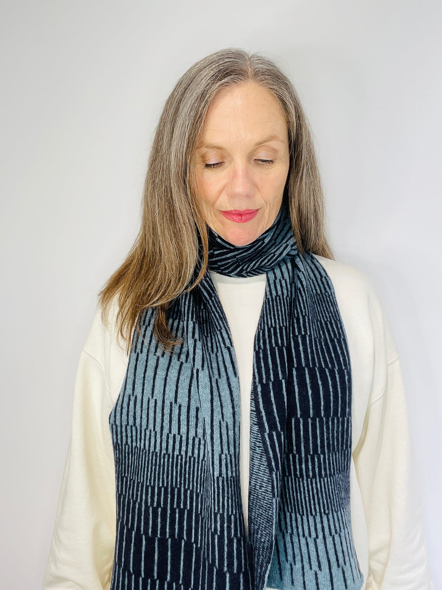 beautiful sexy woman in a white top wearing Blue Caspian and Navy Blue handcrafted merino Lambswool striped Reverb Scarf against a neutral background. Pom Shanty Knitwear