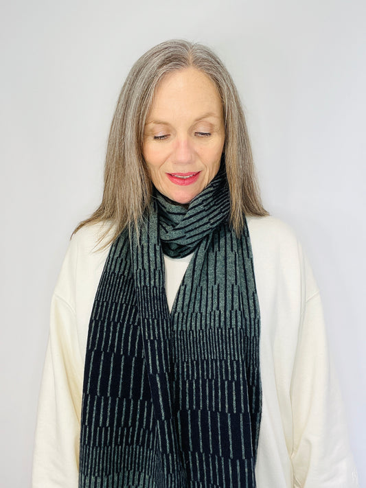 beautiful sexy woman in a white top wearing Peacock Green and Navy Blue handcrafted merino Lambswool striped Reverb Scarf against a neutral background. Pom Shanty Knitwear