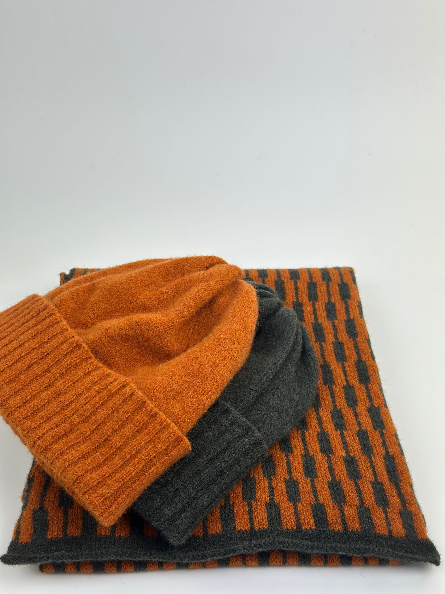 rust and olive green merino lambswool geometric patterned scarf show with  rust and olive green beanies on a plain background.