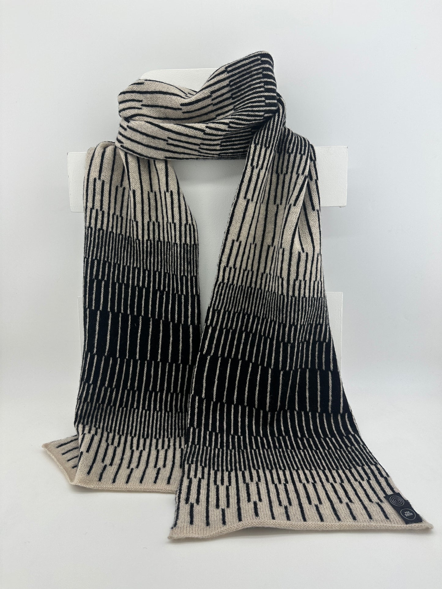Black and Oatmeal handcrafted merino Lambswool striped Reverb Scarf hanging on a white display stand against a neutral background. Pom Shanty Knitwear.