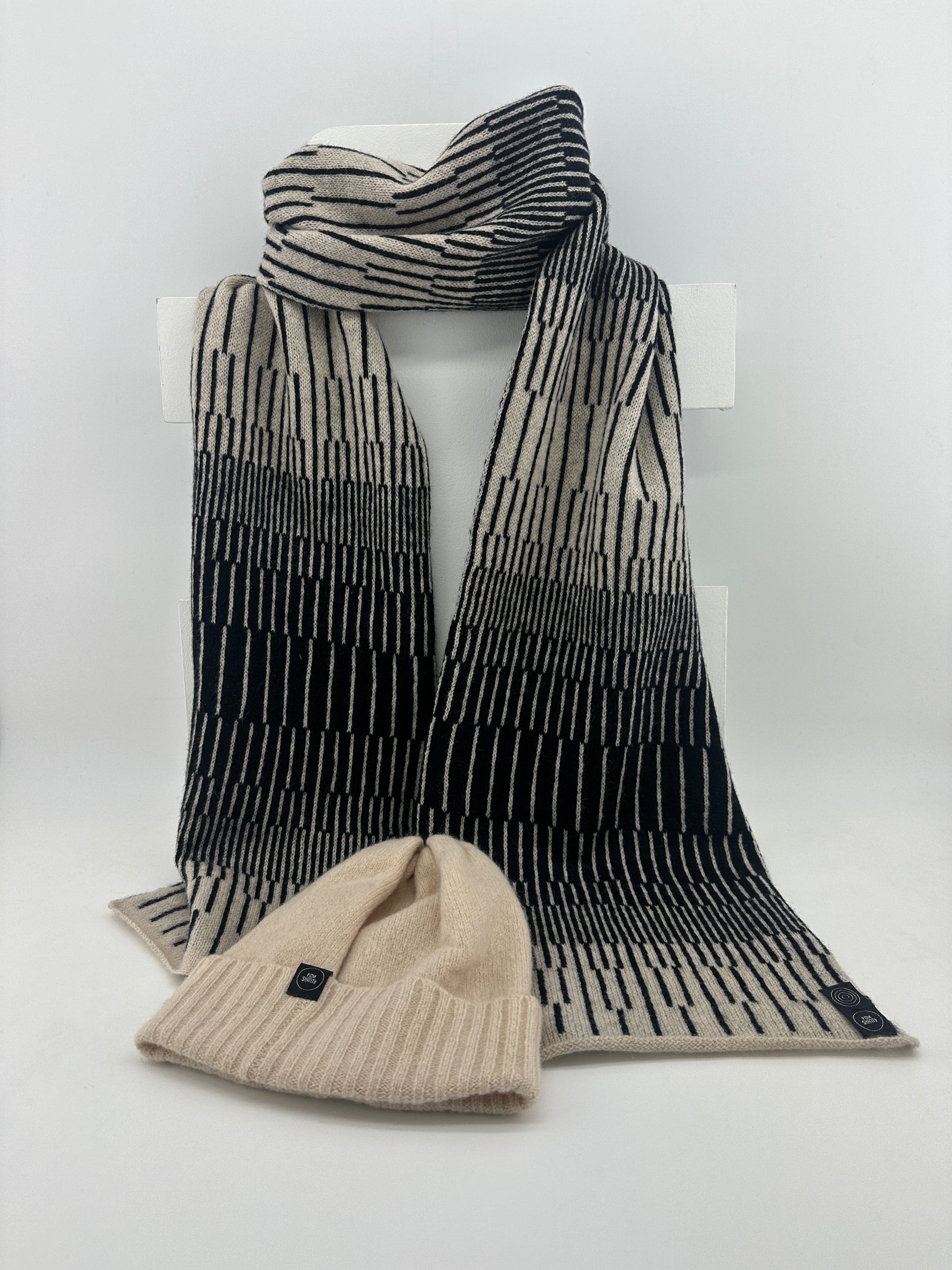 Black and Oatmeal handcrafted merino Lambswool striped Reverb Scarf hanging on a white display stand  with an oatmeal merino lambswool beanie on top of it, against a neutral background. Pom Shanty Knitwear.