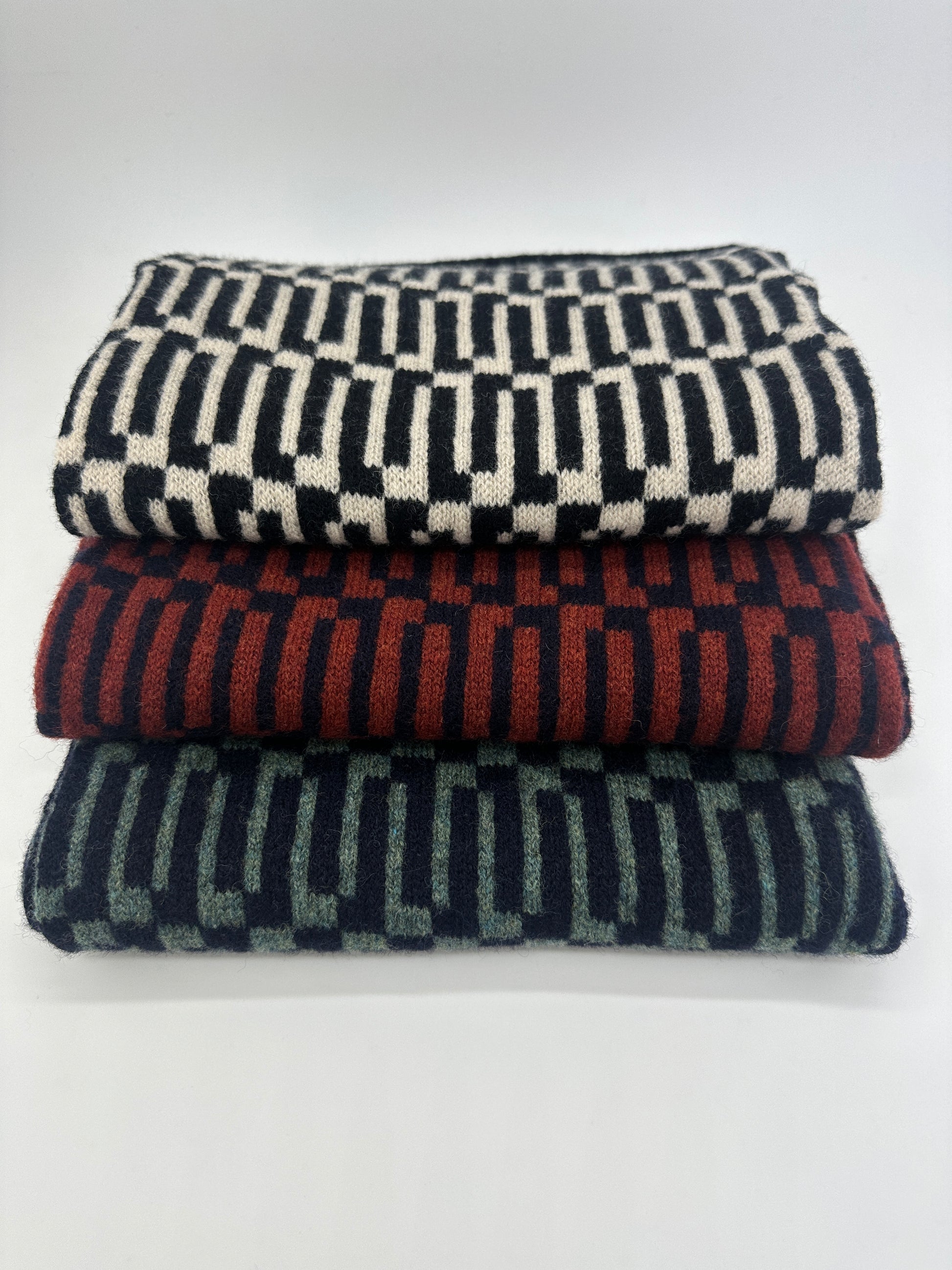 Stack of three different coloured handcrafted merino lambswool Link scarves from Pom Shanty Knitwear on a white table and background