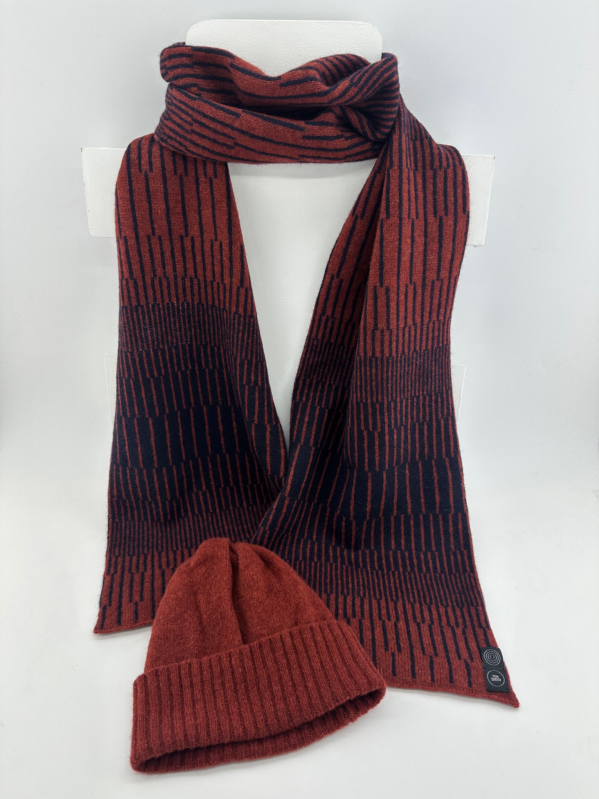 Sienna Brown and Navy Blue handcrafted merino Lambswool striped Reverb Scarf hanging on a white display stand with a sienna brown merino lambswool beanie on top of it, against a neutral background. Pom Shanty Knitwear.