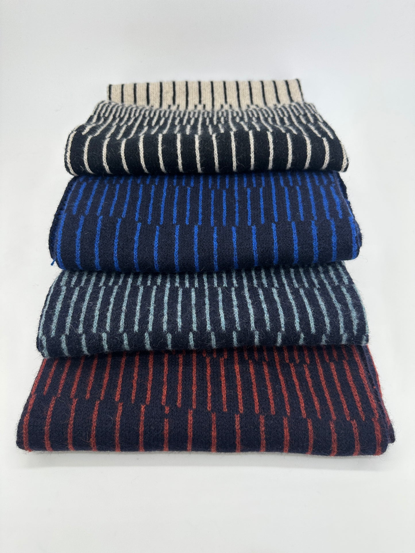 Four different coloured handcrafted merino lambswool striped Reverb scarves folded and stacked on top of each other on a neutral background. Pom Shanty Knitwear.