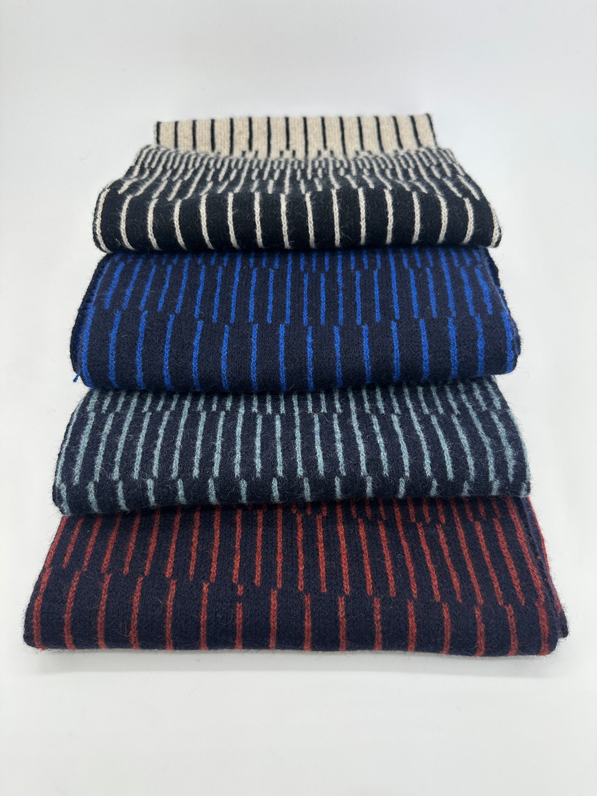 Four different coloured handcrafted merino lambswool striped Reverb scarves folded and stacked on top of each other on a neutral background. Pom Shanty Knitwear.