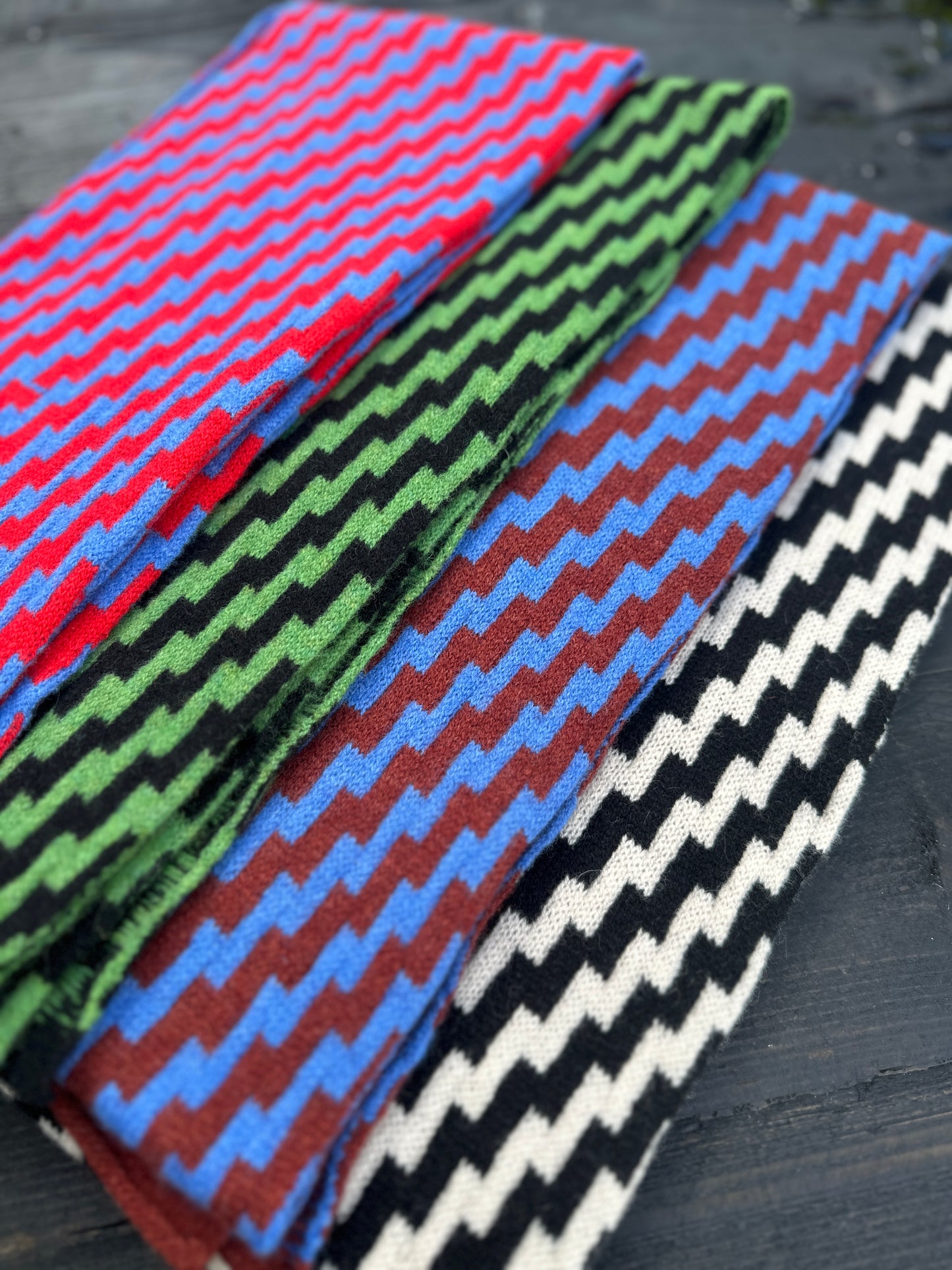 Four rick rack geometric patterned merino lambswool scarves folded and laying spread out on a grey wooden table. pom shanty knitwear