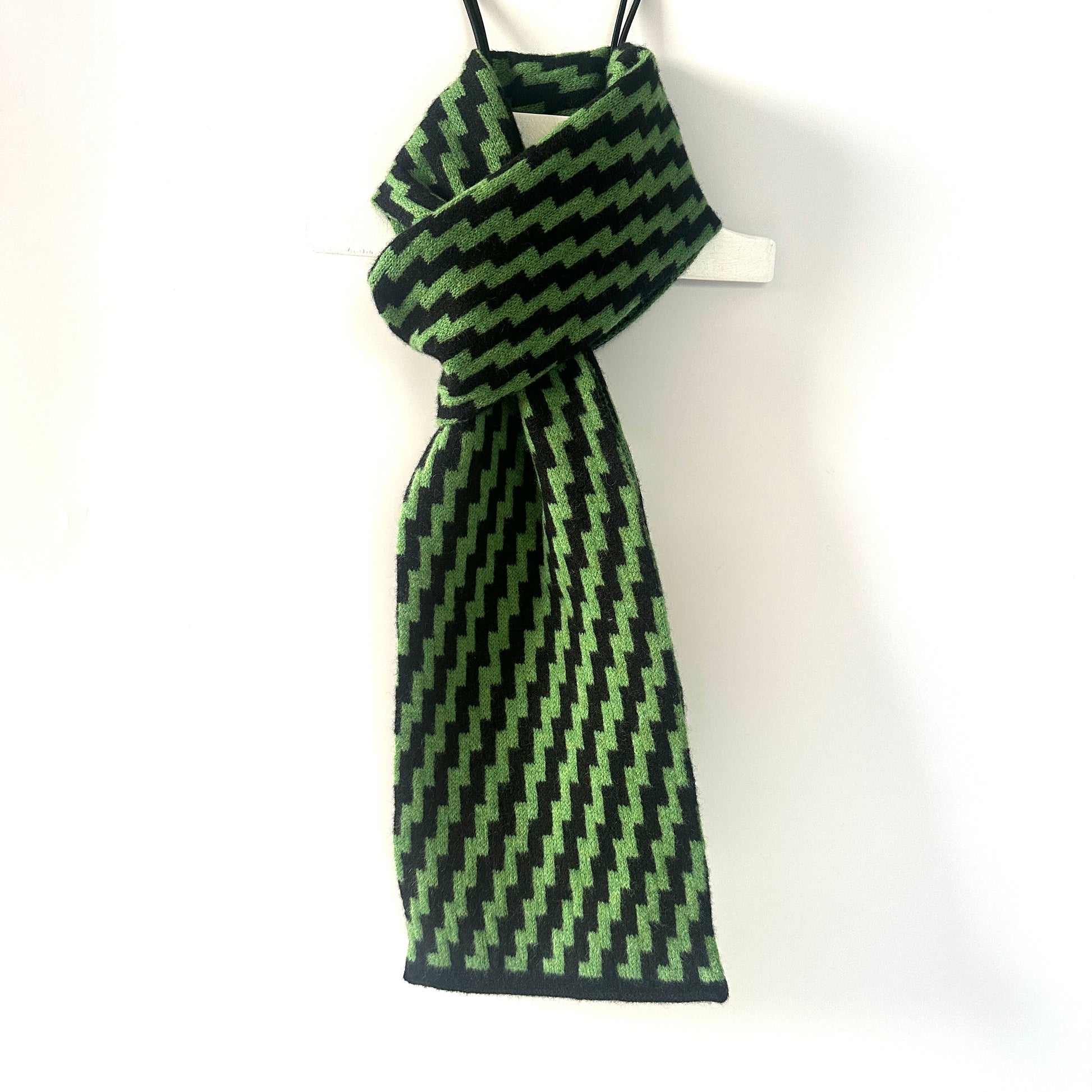 rick rack geometric patterned watercress green and black merino lambswool scarf hanging on a white hanger against a neutral background. Pom Shanty Knitwear.
