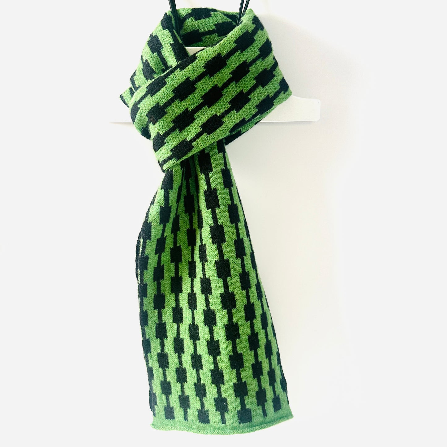 Watercress green and black handcrafted Merino Lambswool geometric patterned Pulse scarf on a white hanger against a white background. Pom Shanty Knitwear.