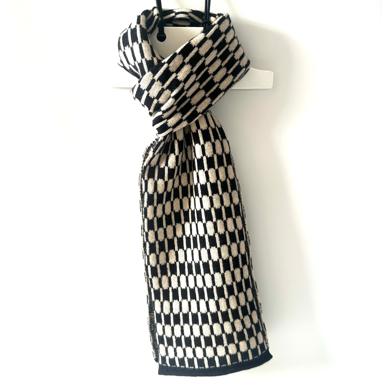 Black and oatmeal handcrafted Merino Lambswool geometric patterned Morse scarf on a white hanger against a white background. Pom Shanty Knitwear.