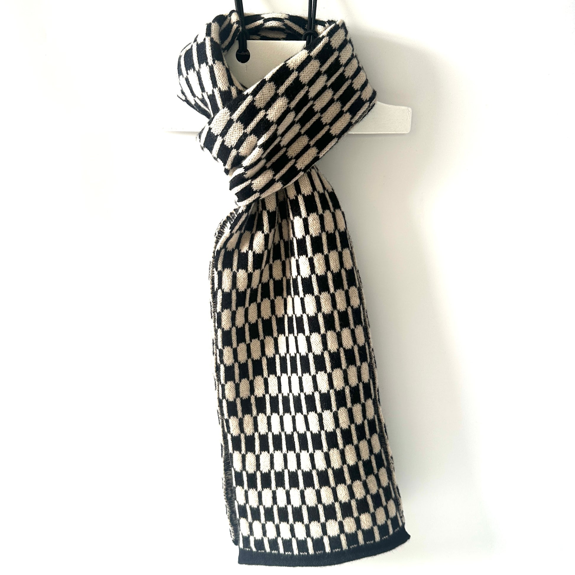 Black and oatmeal handcrafted Merino Lambswool geometric patterned Morse scarf on a white hanger against a white background.  Pom Shanty Knitwear.