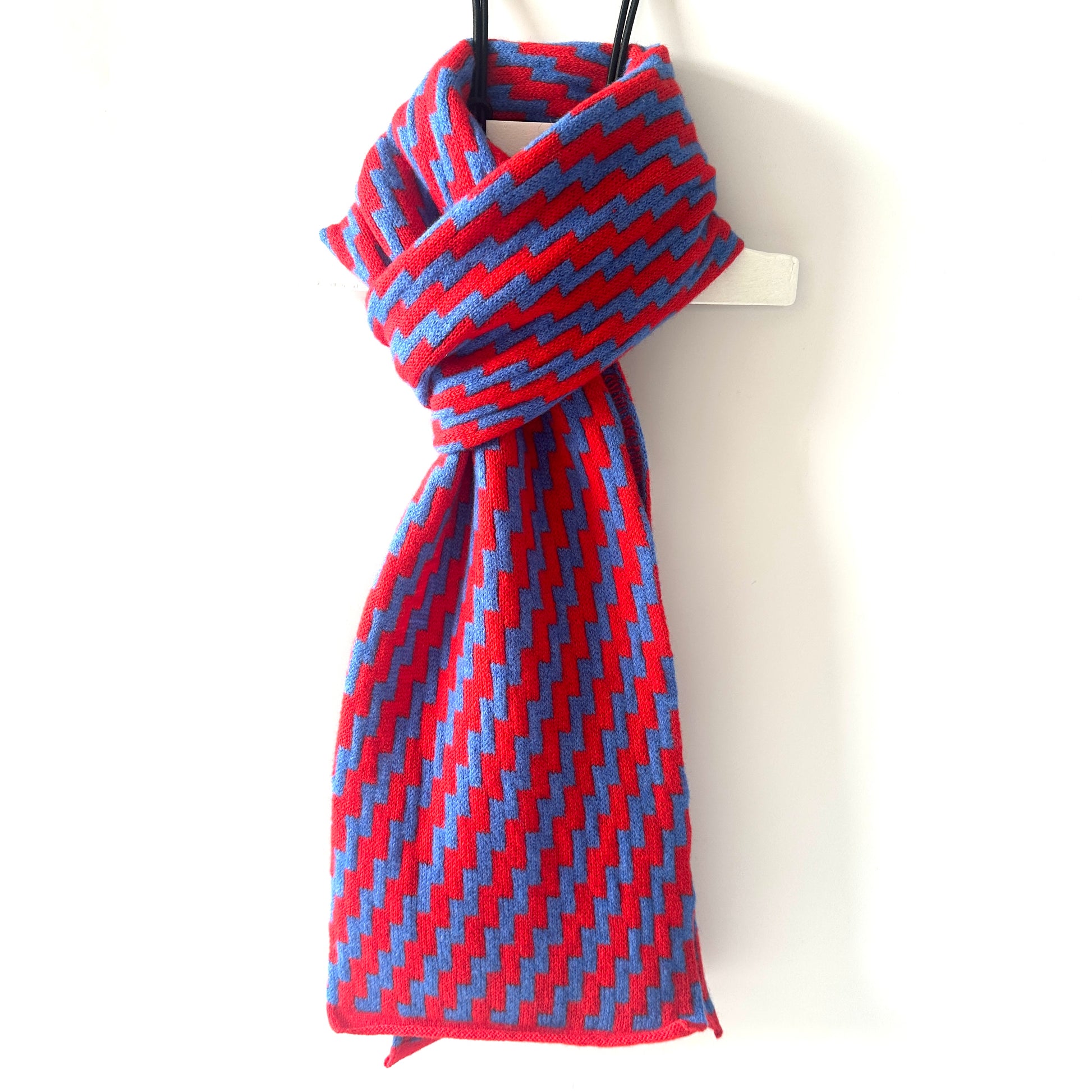 rick rack geometric patterned red and  cornflower blue merino lambswool scarf hanging on a white hanger against a white background. Pom Shanty Knitwear.