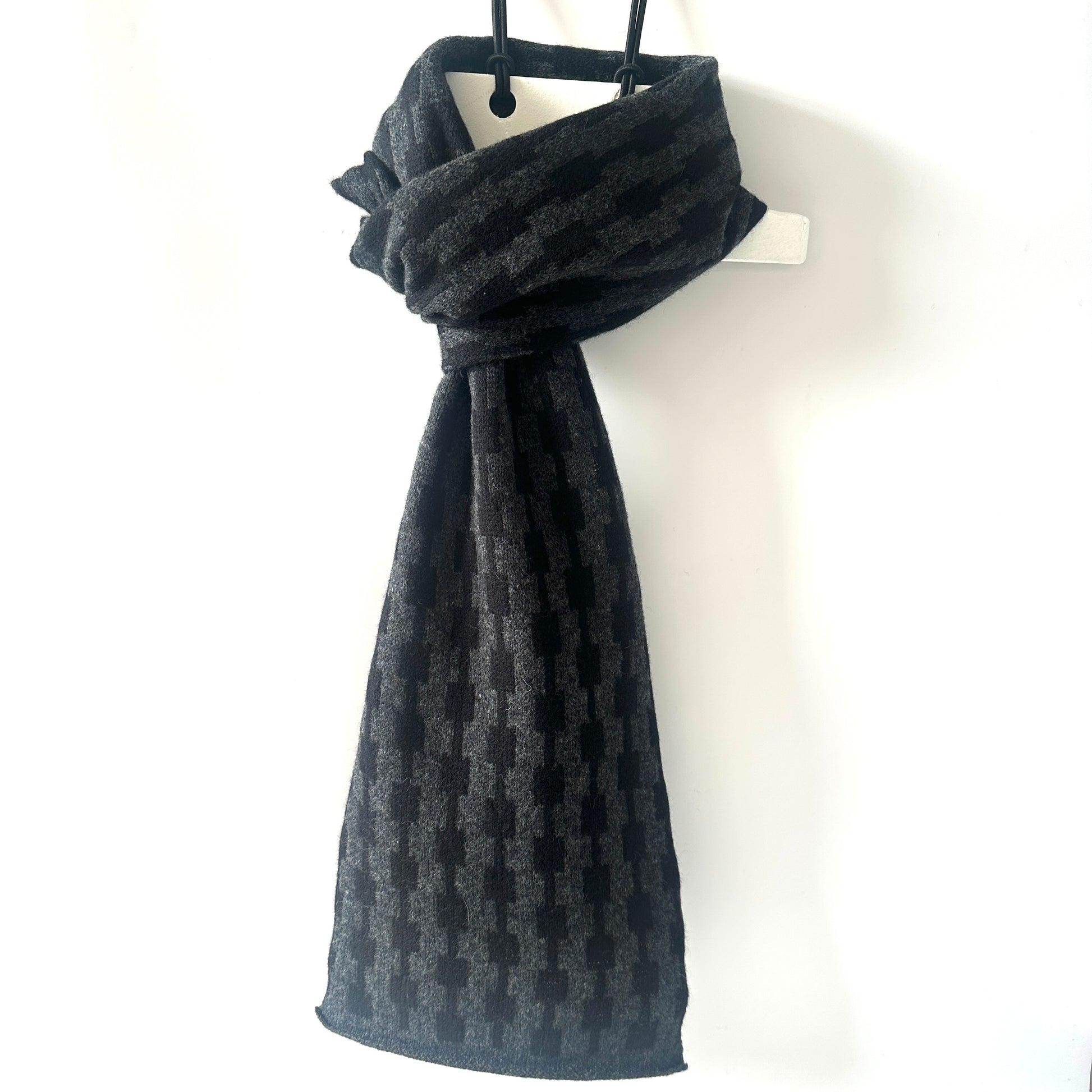 Black and charcoal grey handcrafted Merino Lambswool geometric patterned Pulse scarf on a white hanger against a white background.  Pom Shanty Knitwear.