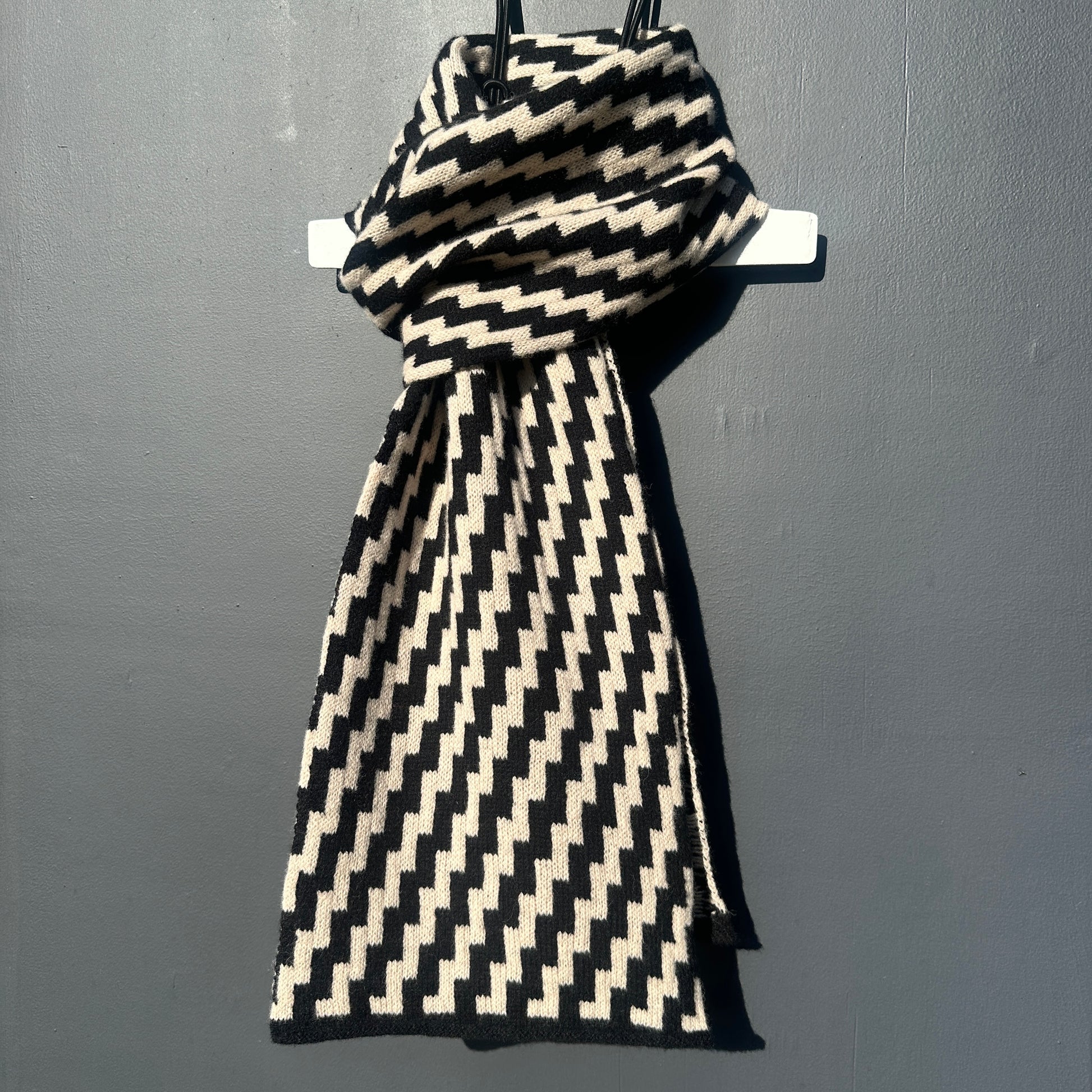 rick rack geometric patterned black and oatmeal merino lambswool scarf tied on a hanger on a rod against a grey background. Pom Shanty Knitwear.