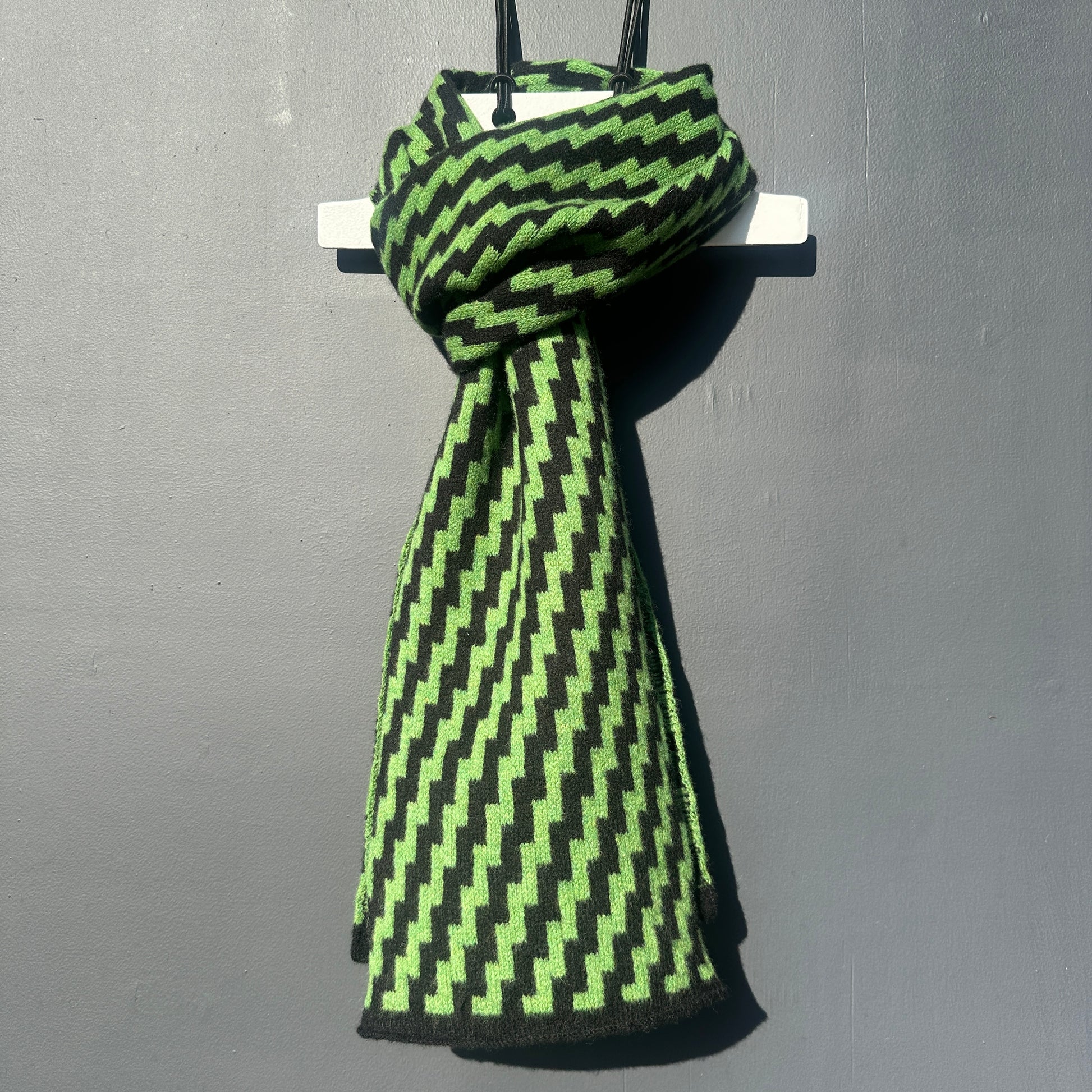 rick rack geometric patterned watercress green and black merino lambswool scarf hanging on a rod against a grey background. Pom Shanty Knitwear.