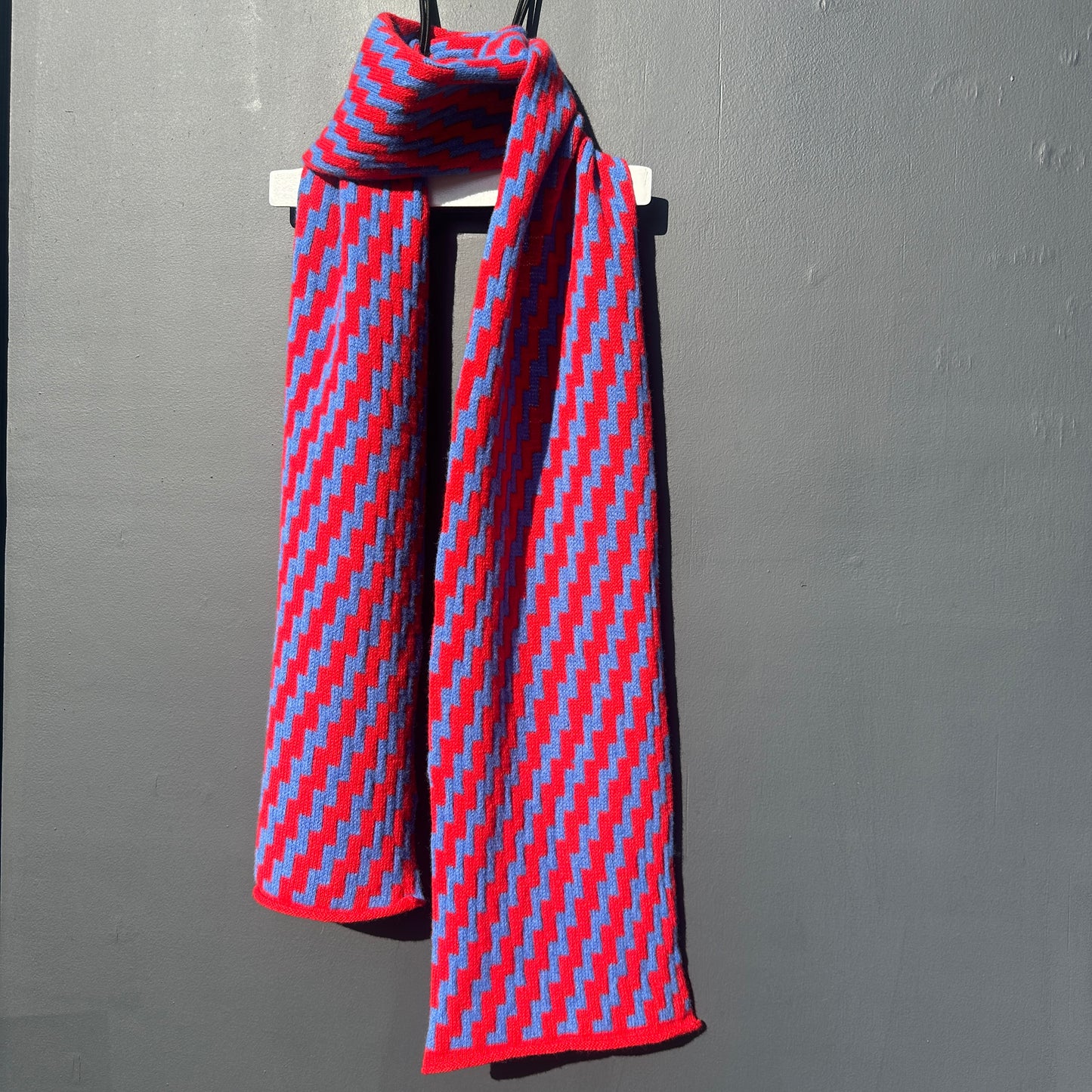 rick rack geometric patterned red and cornflower blue merino lambswool scarf draped on a white hanger against a grey background. Pom Shanty Knitwear.