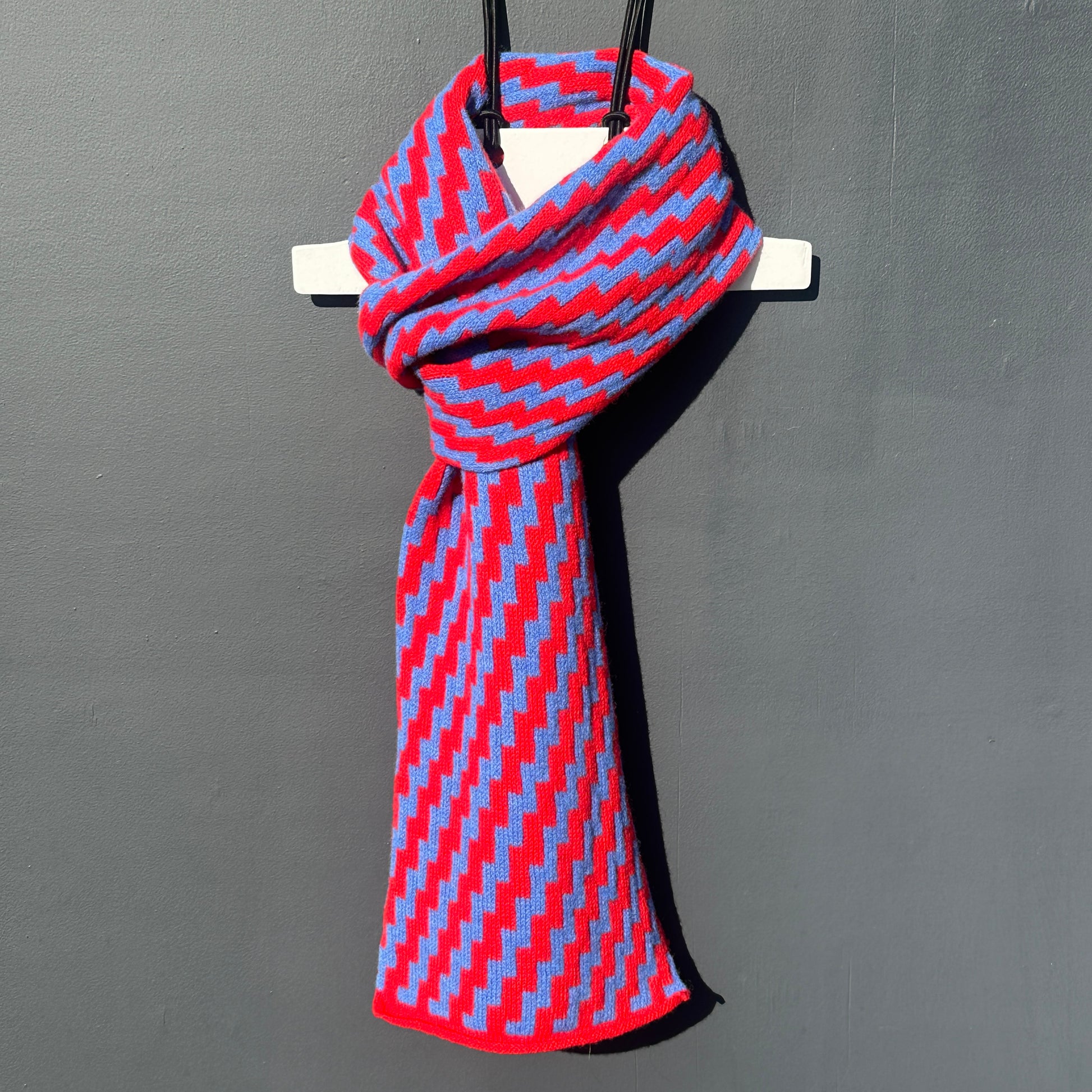 rick rack geometric patterned red and  cornflower blue merino lambswool scarf hanging on a white hanger against a grey background. Pom Shanty Knitwear.