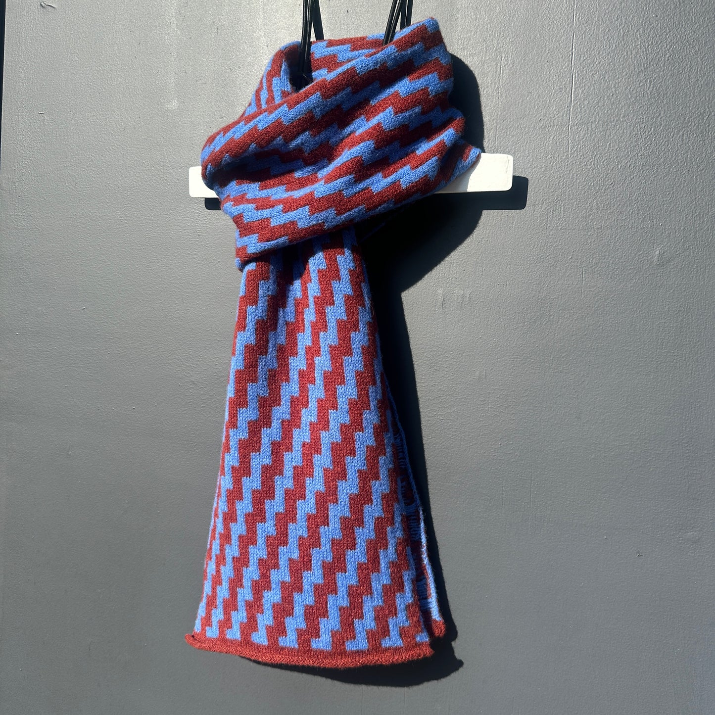rick rack geometric patterned sienna brown and cornflower blue merino lambswool scarf hanging on a white hanger against a grey background. Pom Shanty Knitwear.