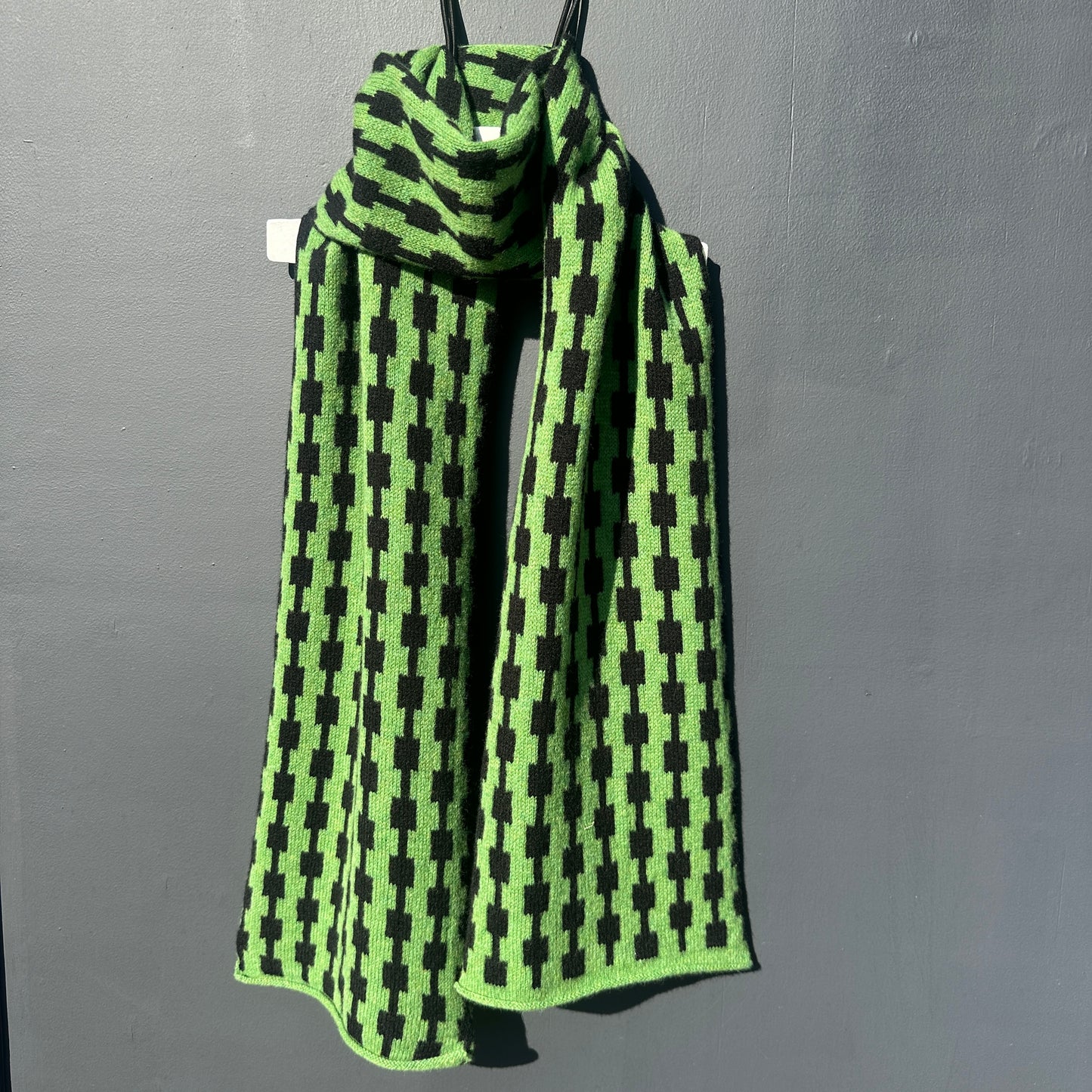 Watercress green and black handcrafted Merino Lambswool geometric patterned Pulse scarf on a white hanger against a grey background. Different drapping. Pom Shanty Knitwear.
