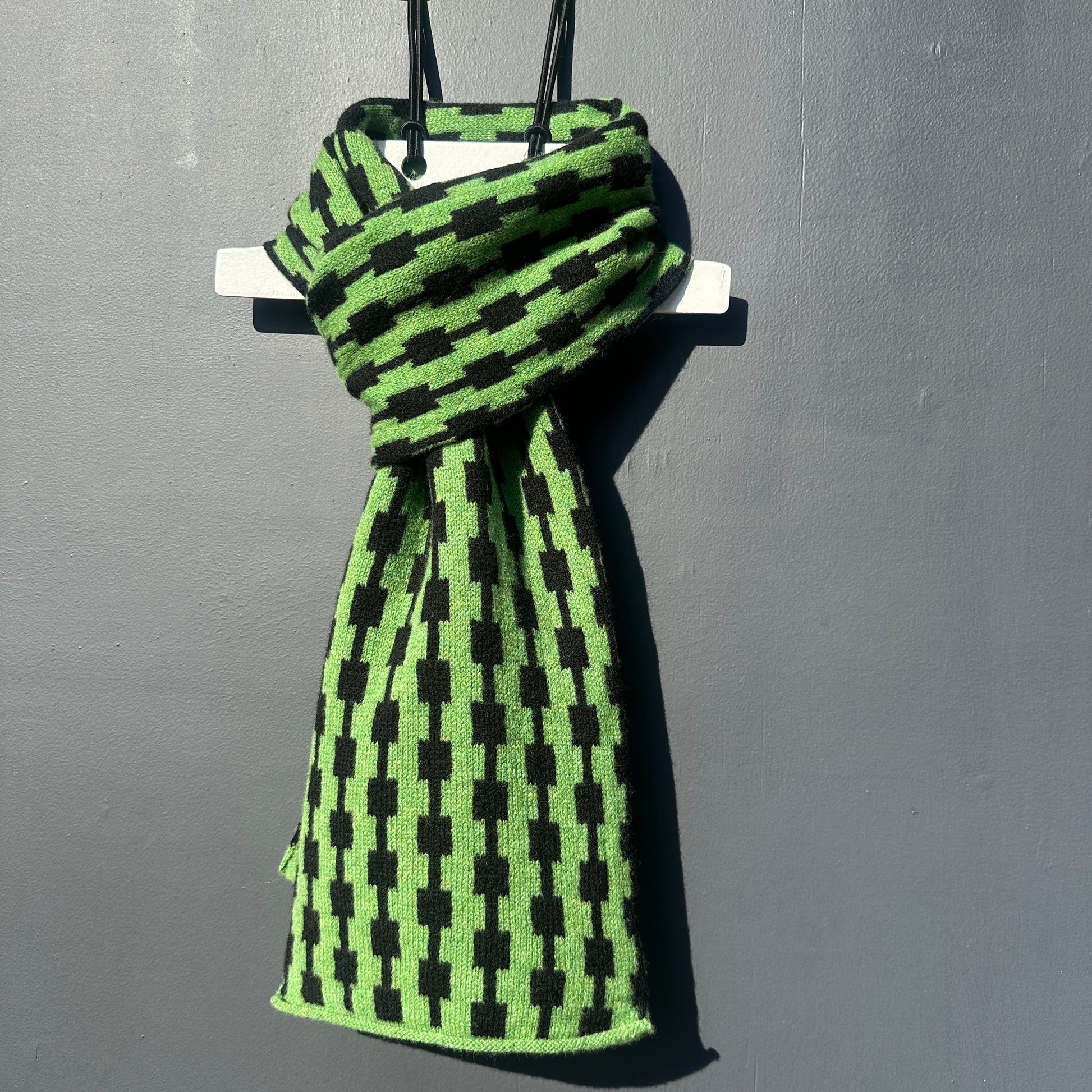 Watercress green and black handcrafted Merino Lambswool geometric patterned Pulse scarf on a white hanger against a grey background.  Pom Shanty Knitwear.