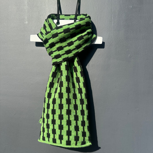 Watercress green and black handcrafted Merino Lambswool geometric patterned Pulse scarf on a white hanger against a grey background.  Pom Shanty Knitwear.