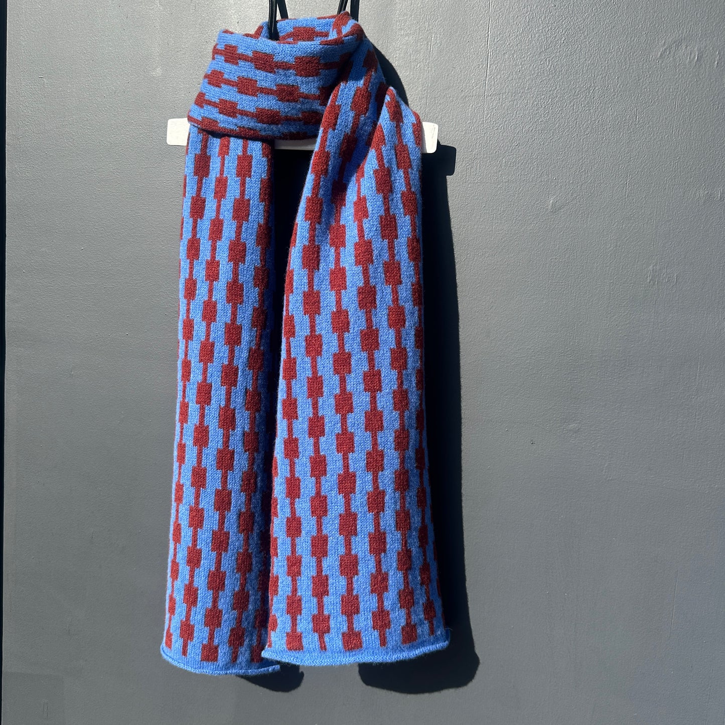 Cornflower blue and sienna brown handcrafted Merino Lambswool geometric patterned Pulse scarf on a white hanger against a grey background. Pom Shanty Knitwear.