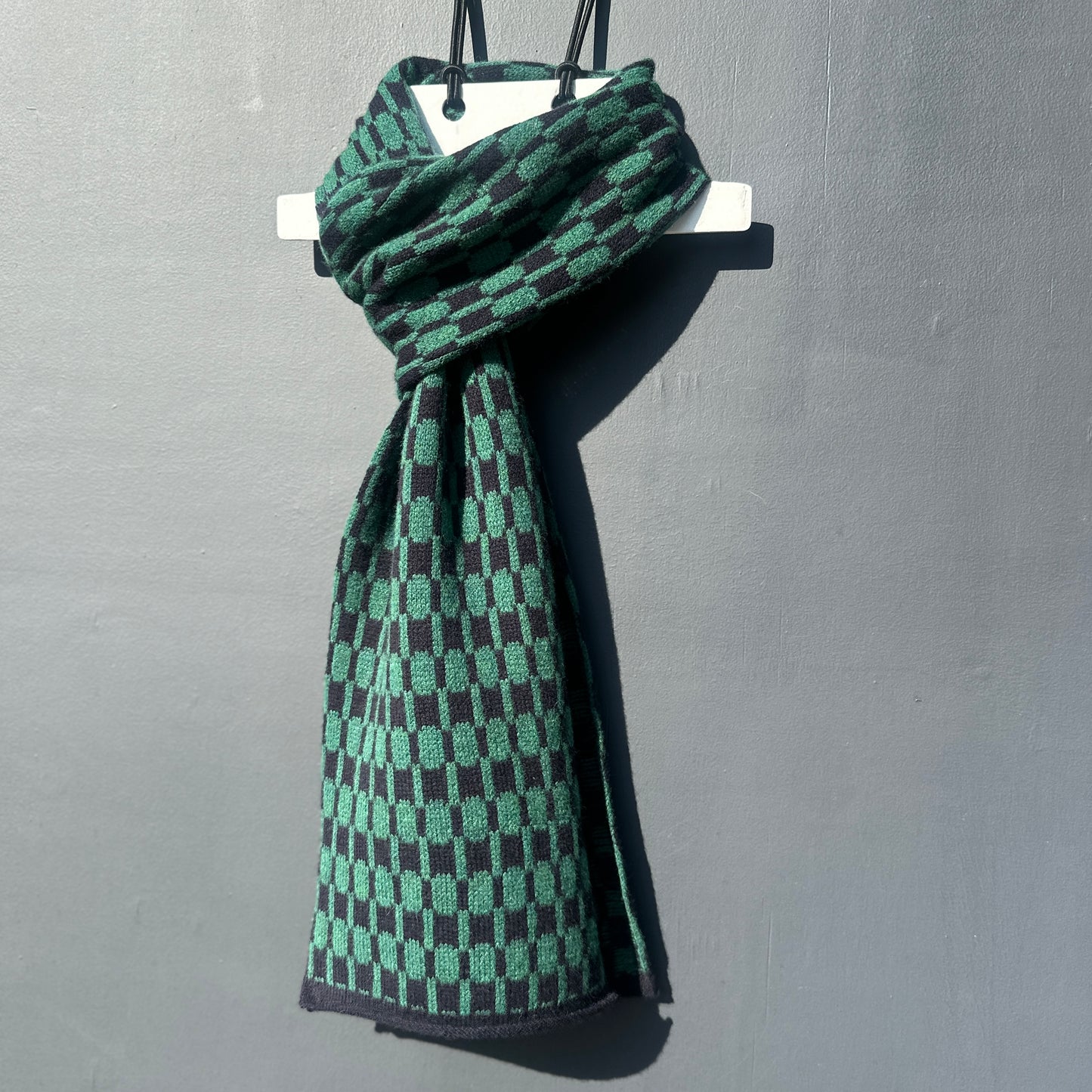 Navy blue and cossack green handcrafted Merino Lambswool geometric patterned Morse scarf on a white hanger against a grey background.  Pom Shanty Knitwear.