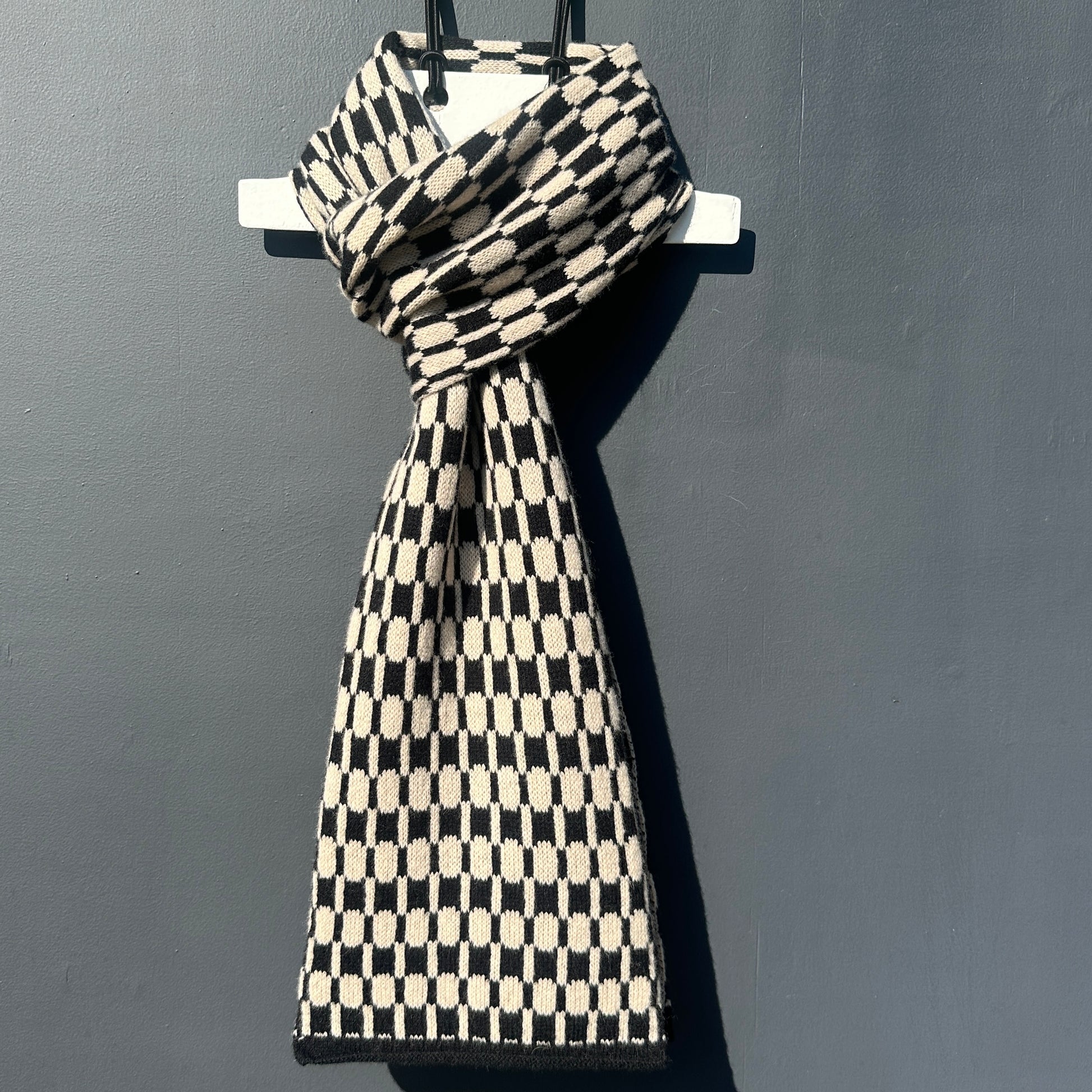 Black and oatmeal handcrafted Merino Lambswool geometric patterned Morse scarf on a white hanger against a grey background.  Pom Shanty Knitwear.