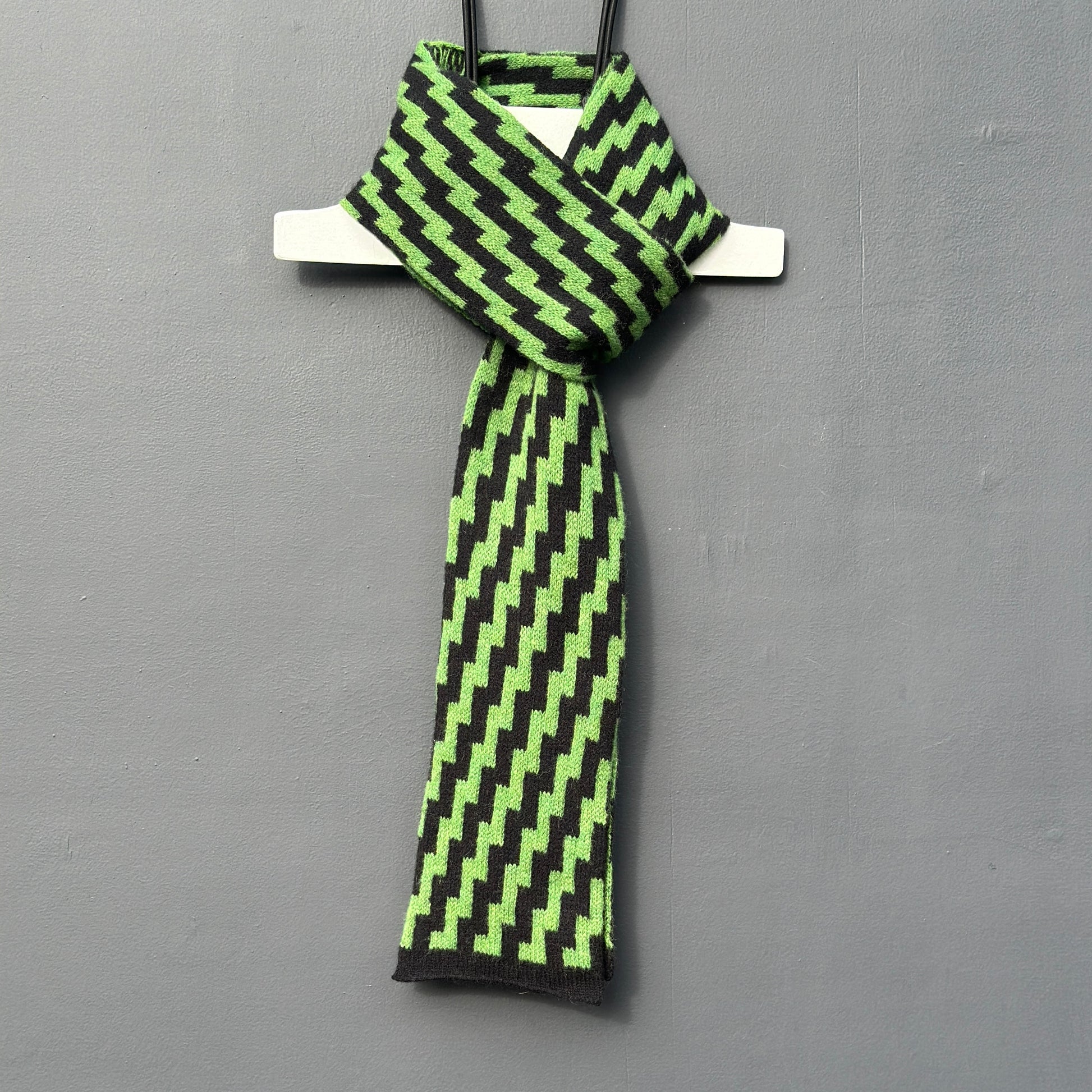 rick rack geometric patterned watercress green and black merino lambswool skinny scarf hanging on a rod against a grey background. Pom Shanty Knitwear.