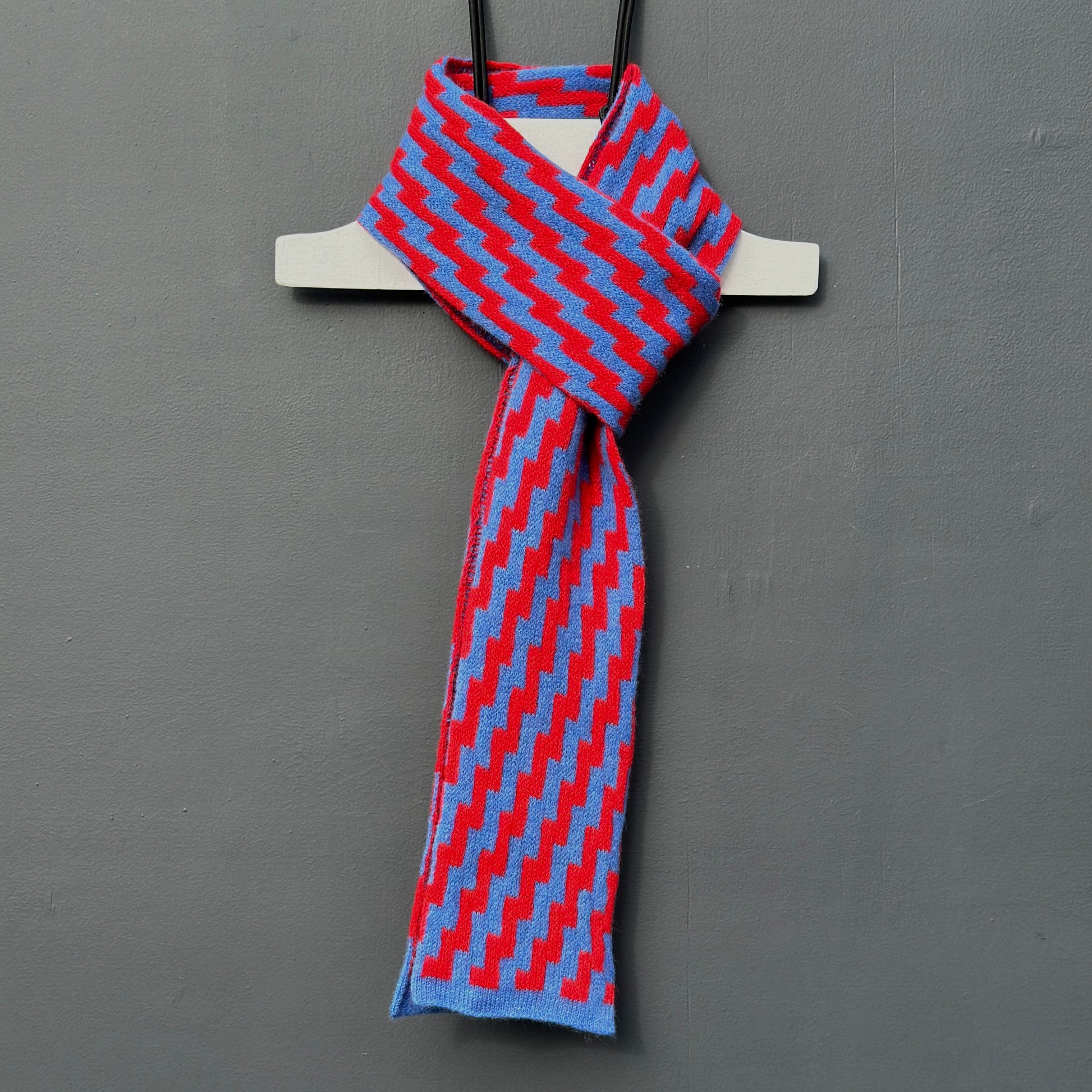 rick rack geometric patterned red and  cornflower blue merino lambswool skinny scarf hanging on a rod against a grey background. Pom Shanty Knitwear.