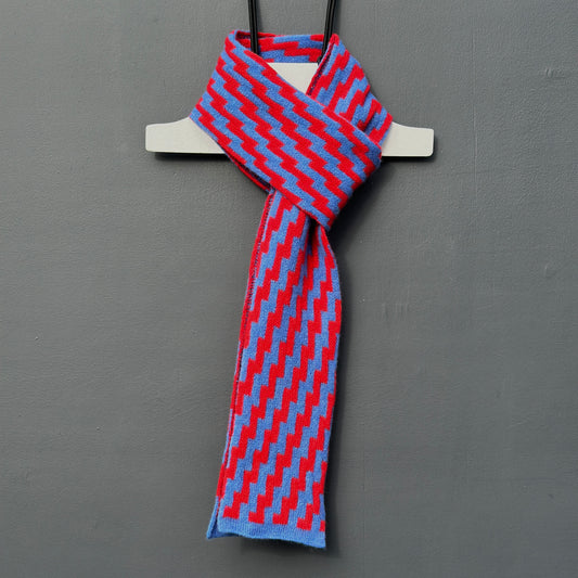 rick rack geometric patterned red and  cornflower blue merino lambswool skinny scarf hanging on a rod against a grey background. Pom Shanty Knitwear.