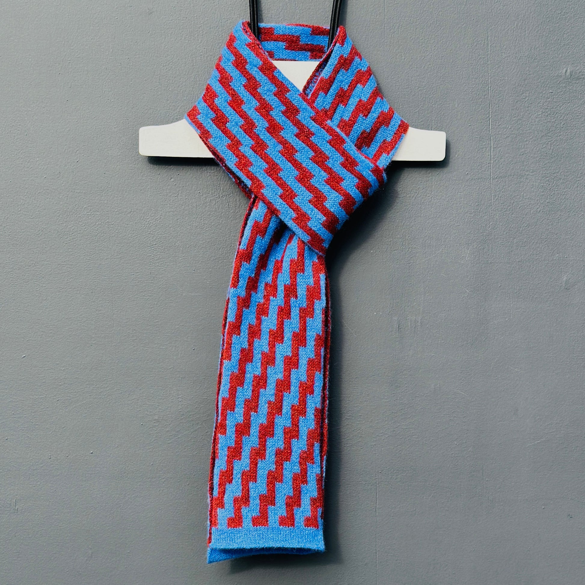 rick rack geometric patterned cornflower blue and sienna brown merino lambswool skinny scarf tied on a hanger against a grey background