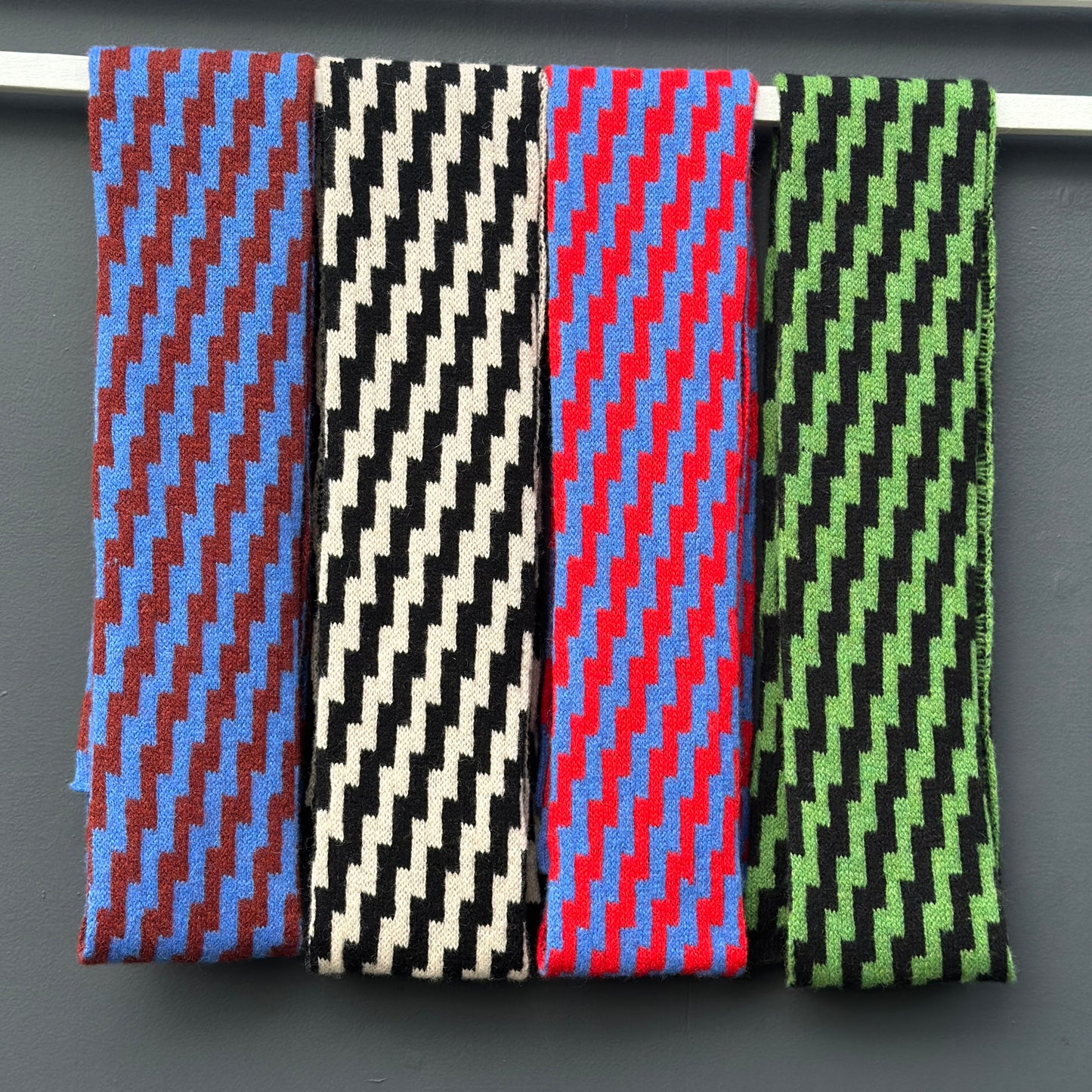 four rick rack geometric patterned merino lambswool skinny scarves hanging on a rod against a grey background