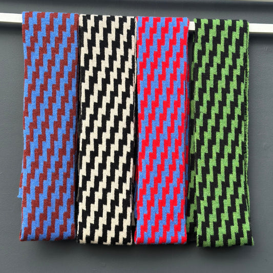 Four rick rack geometric patterned merino lambswool skinny scarves hanging on a rod against a grey background. pom shanty knitwear