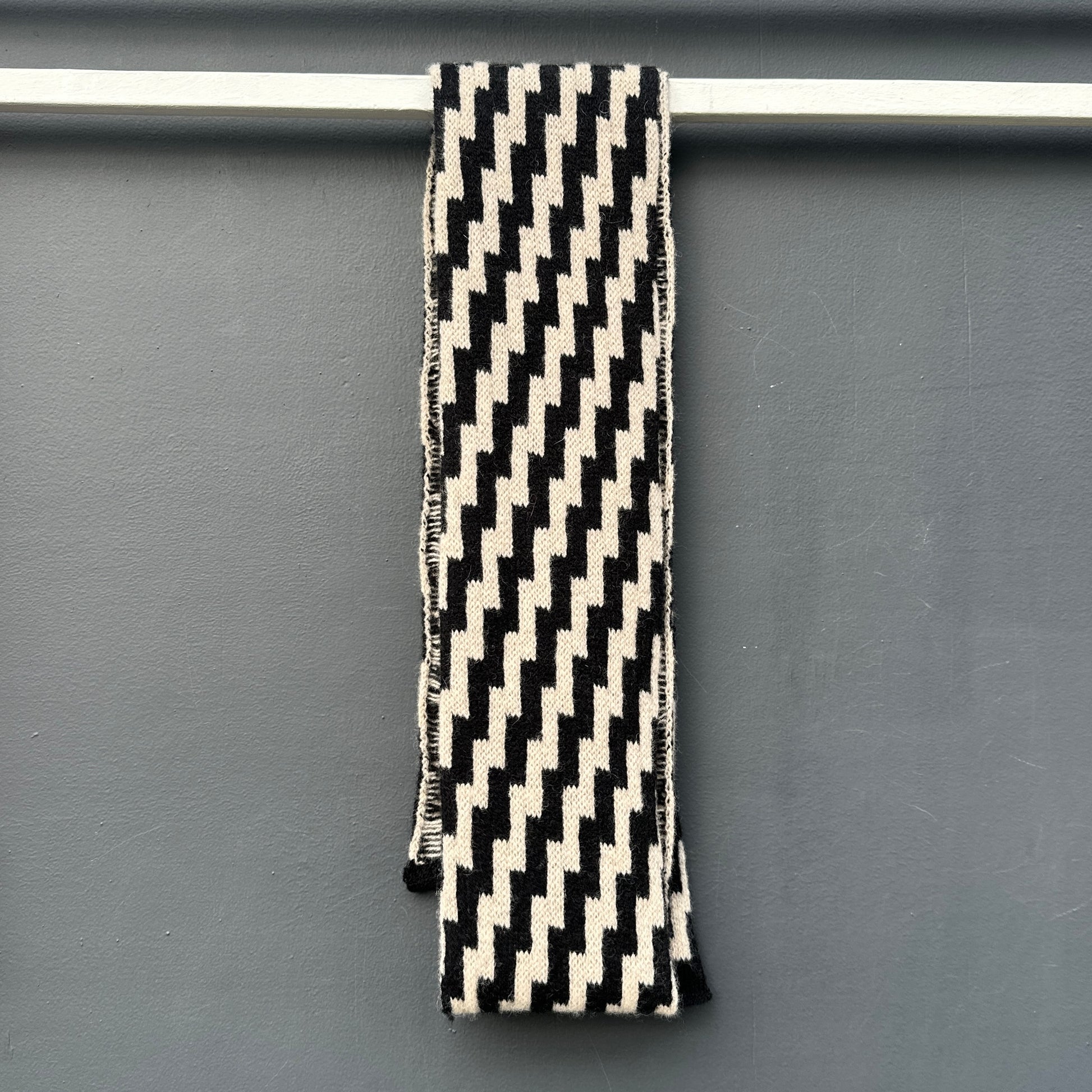 rick rack geometric patterned black and oatmeal merino lambswool skinny scarf hanging on a rod against a grey background