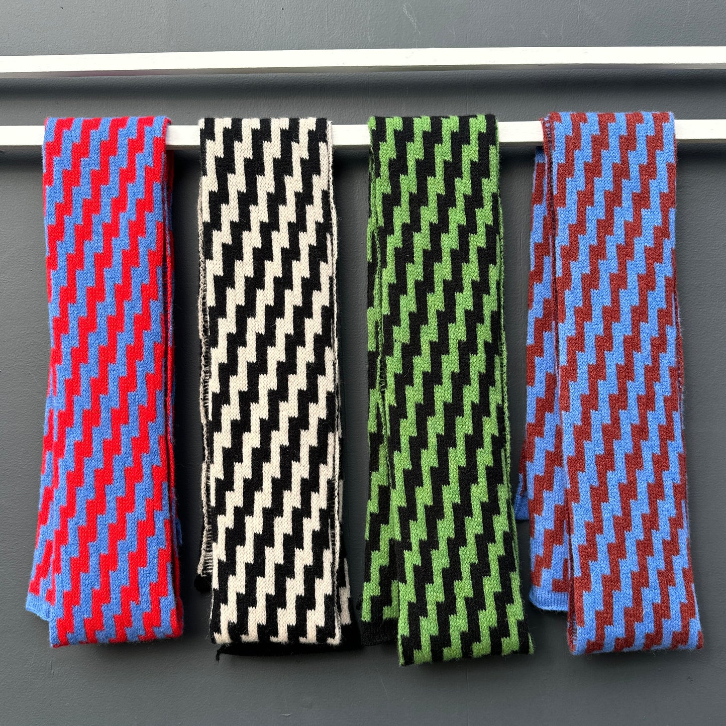 Four rick rack geometric patterned merino lambswool skinny scarves hanging on a white rod against a grey background. Pom Shanty Knitwear.