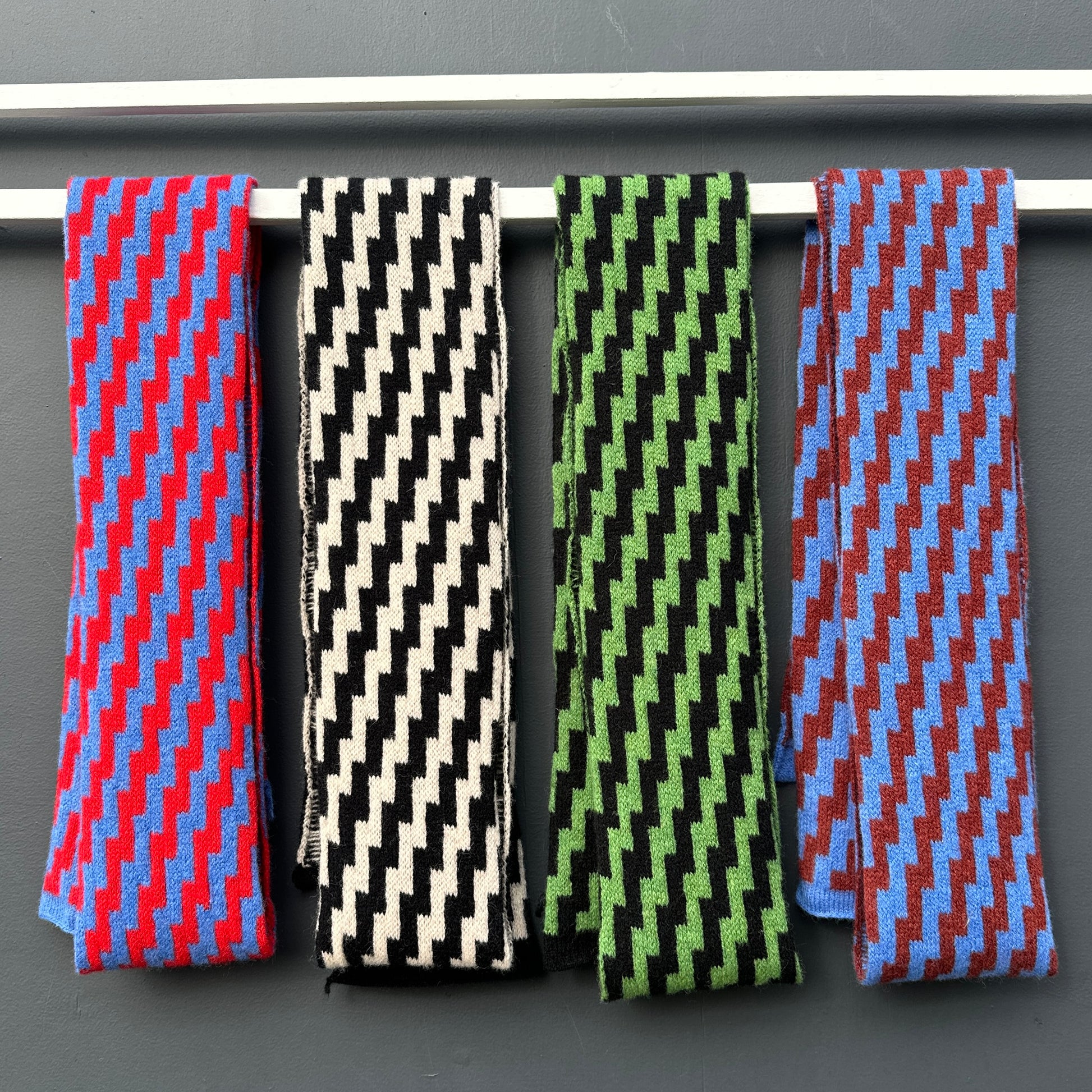 Four rick rack geometric patterned merino lambswool skinny scarves hanging on a white rod against a grey background. Pom Shanty Knitwear.