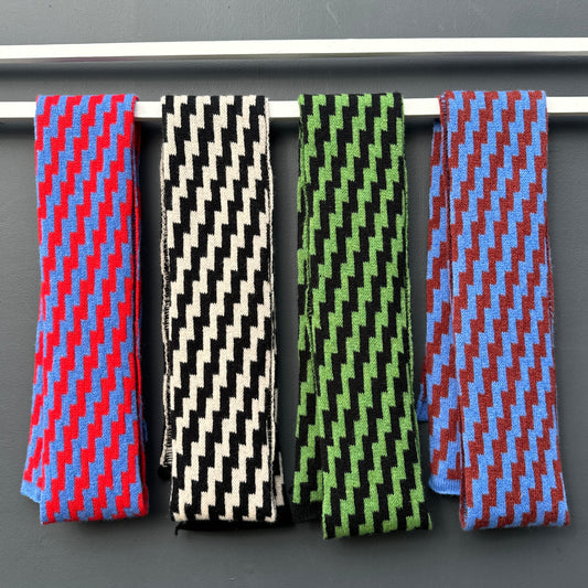 Four rick rack geometric patterned merino lambswool skinny scarves hanging on a white rod against a grey background. Pom Shanty Knitwear.