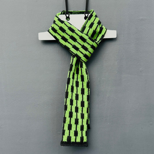 geometric patterned watercress green and black merino lambswool skinny scarf hanging on a rod against a gray background.