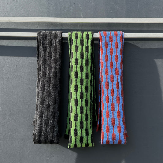 A charcoal grey and black, a watercress green and black and a cornflower blue and sienna brown geometric patterned merino lambswool skinny scarves hanging on a rod against a gray background.