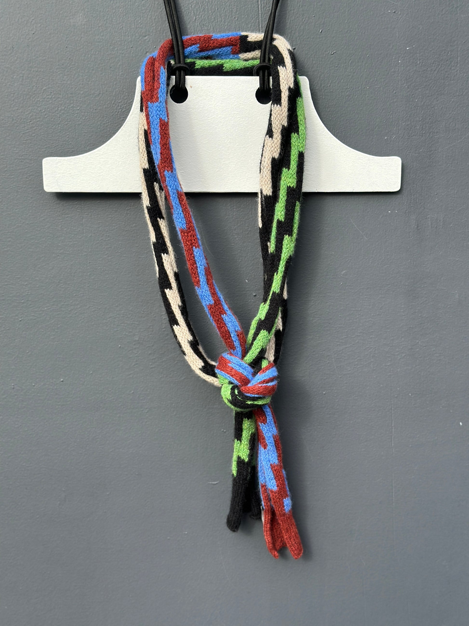 A multi-coloured rick rack patterned Pom Shanty Knitwear merino lambswool neck tie hanging on a white hanger againts a grey background for men, women and unisex.
