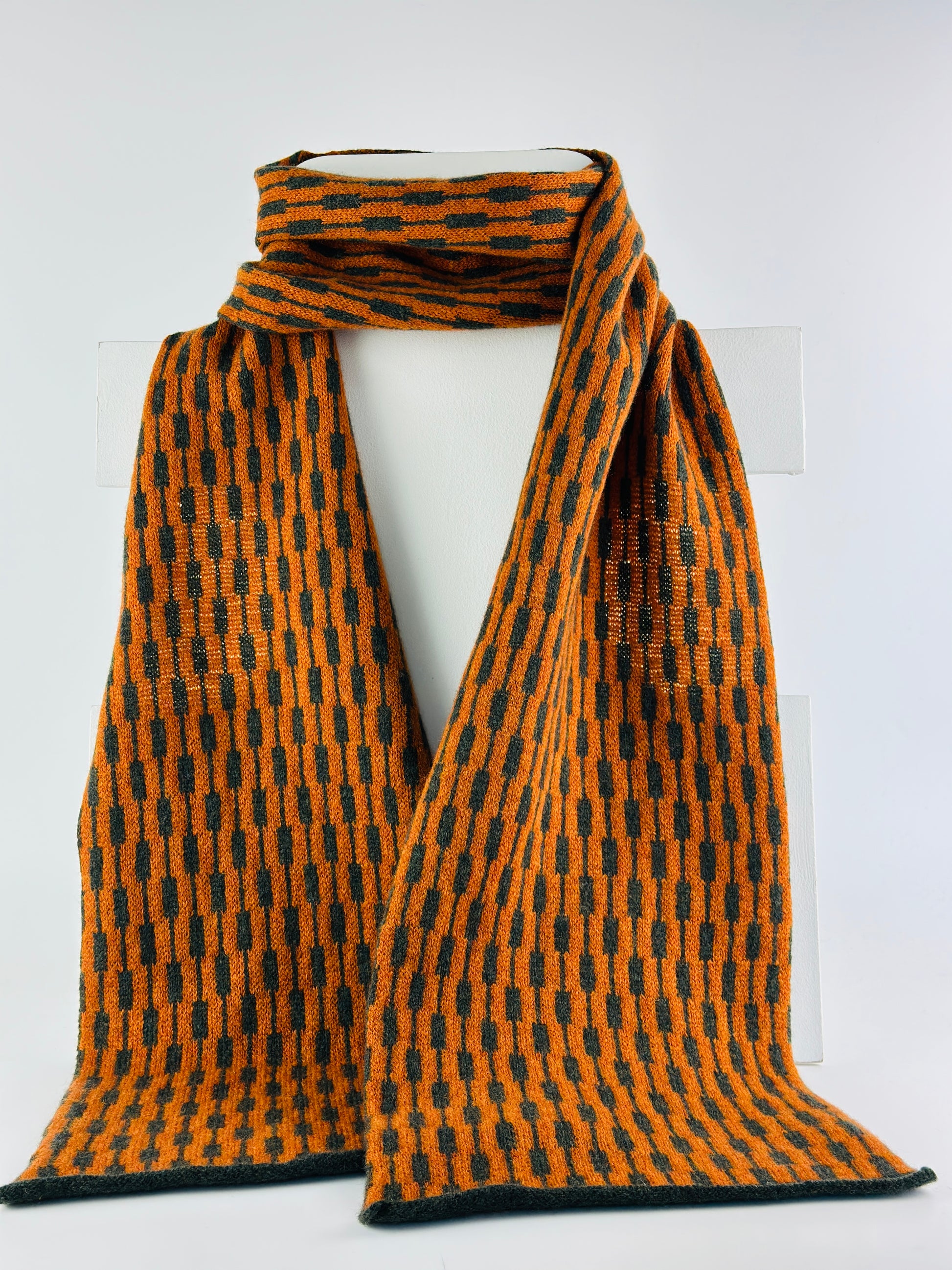 rust and olive green merino lambswool geometric patterned scarf on a rack on a plain background.