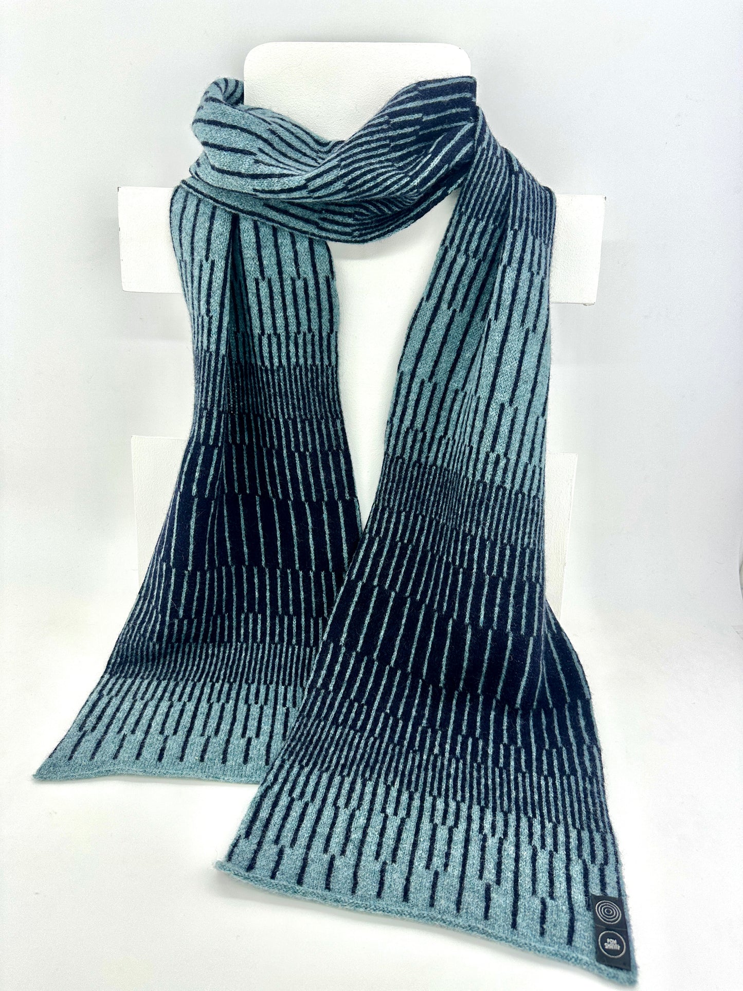Blue Caspian and Navy Blue handcrafted merino Lambswool striped Reverb Scarf hanging on a white display stand  against a neutral background. Pom Shanty Knitwear.