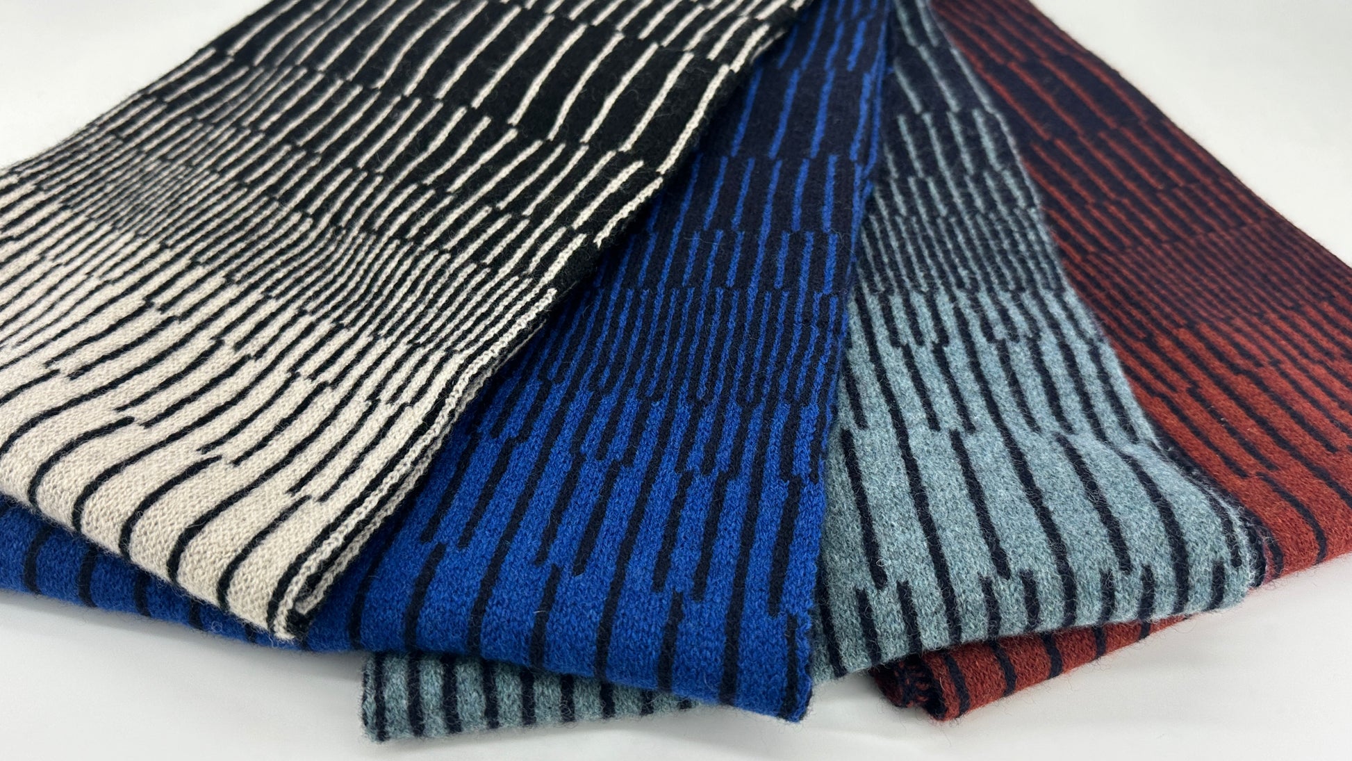 Four different coloured handcrafted merino lambswool striped Reverb scarves folded and fanned out on a white background. Pom Shanty Knitwear.