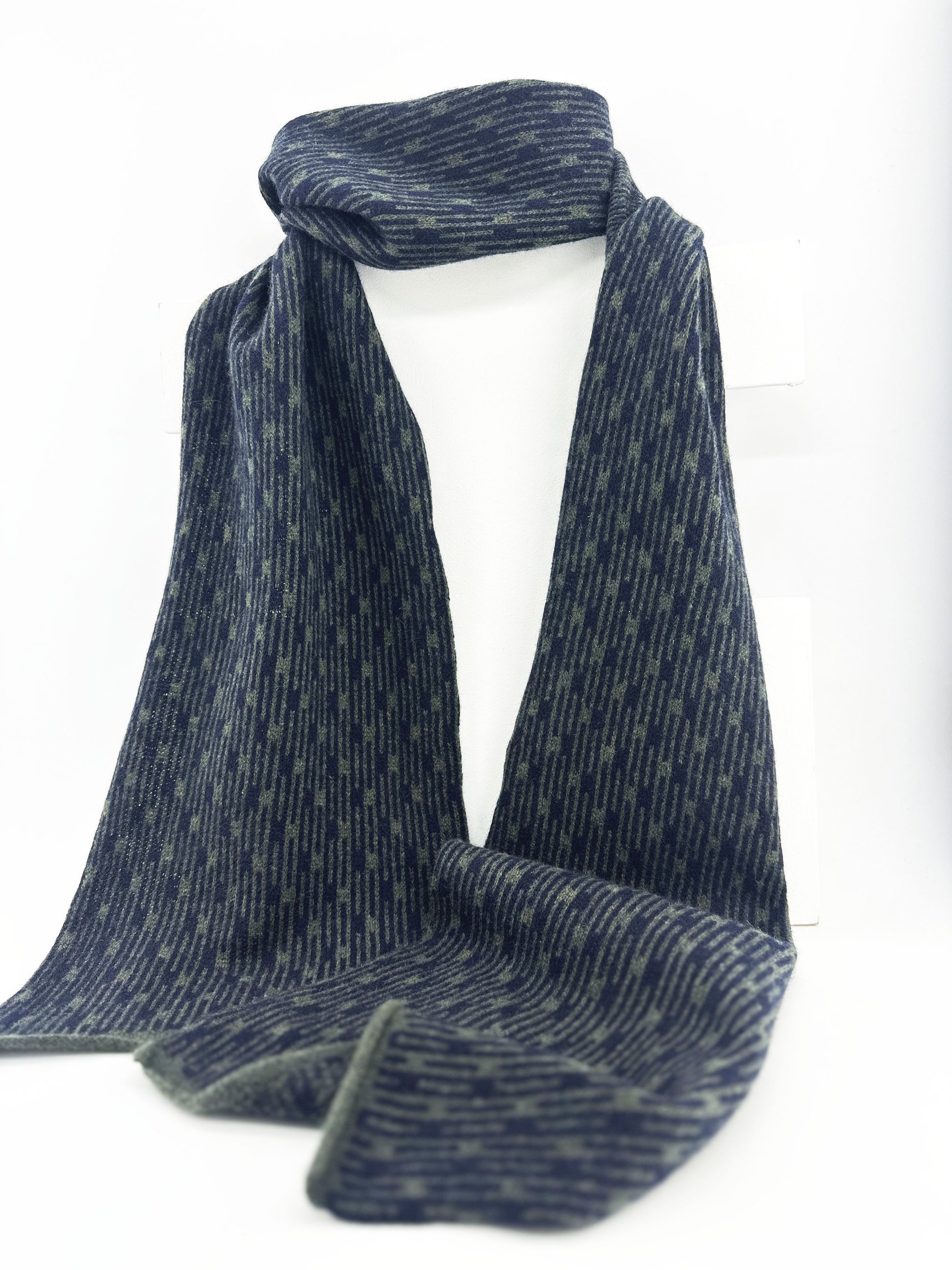 Olive Green and Navy Blue Metropolis geometric patterned handcrafted merino lambswool scarf hanging on a white display stand on a neutral background.  Pom Shanty Knitwear