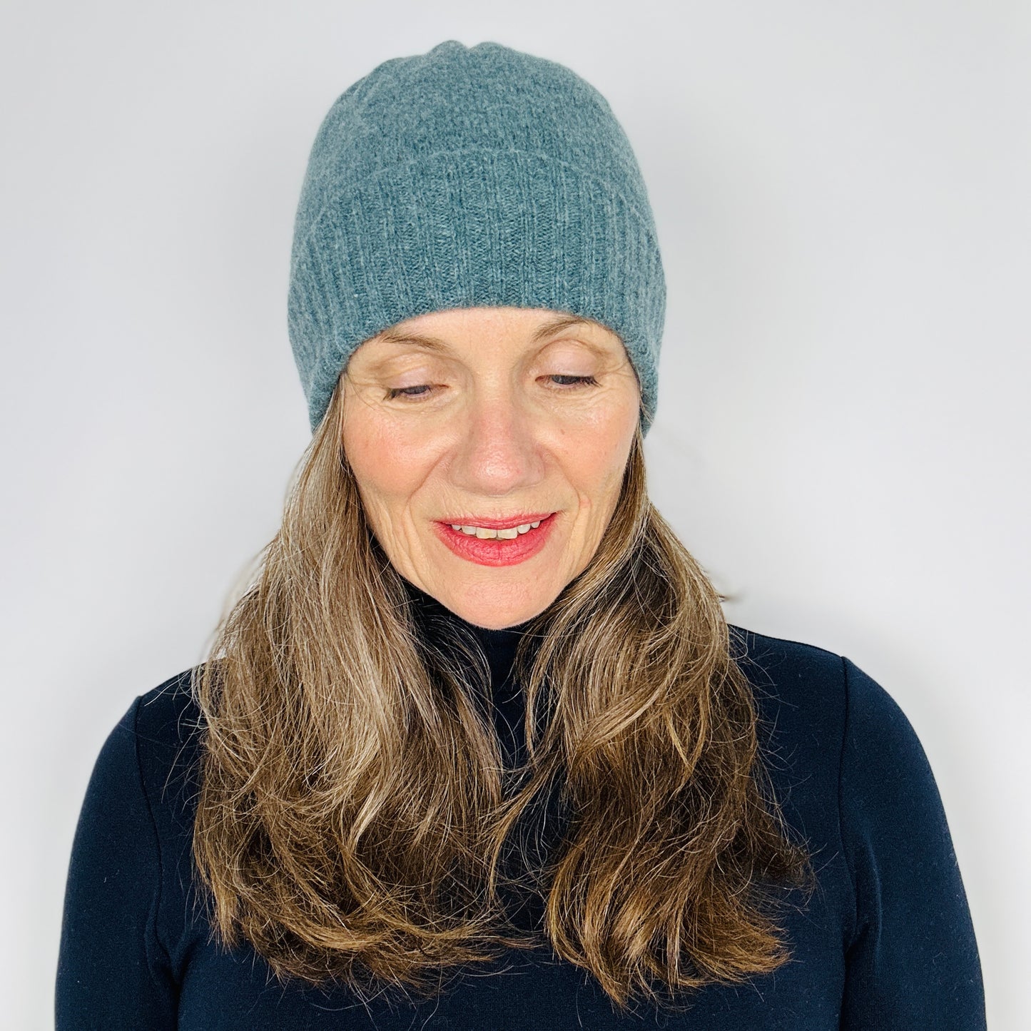 beautiful sexy woman in a black top wearing a caspian blue merino lambswool beanie with a double ribbed brim on a plain background.