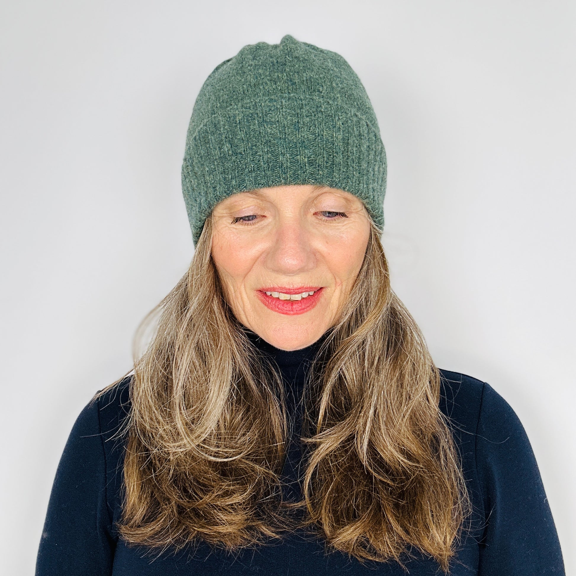 beautiful sexy woman in a black top wearing a peacock green merino lambswool beanie with a double ribbed brim on a plain background.