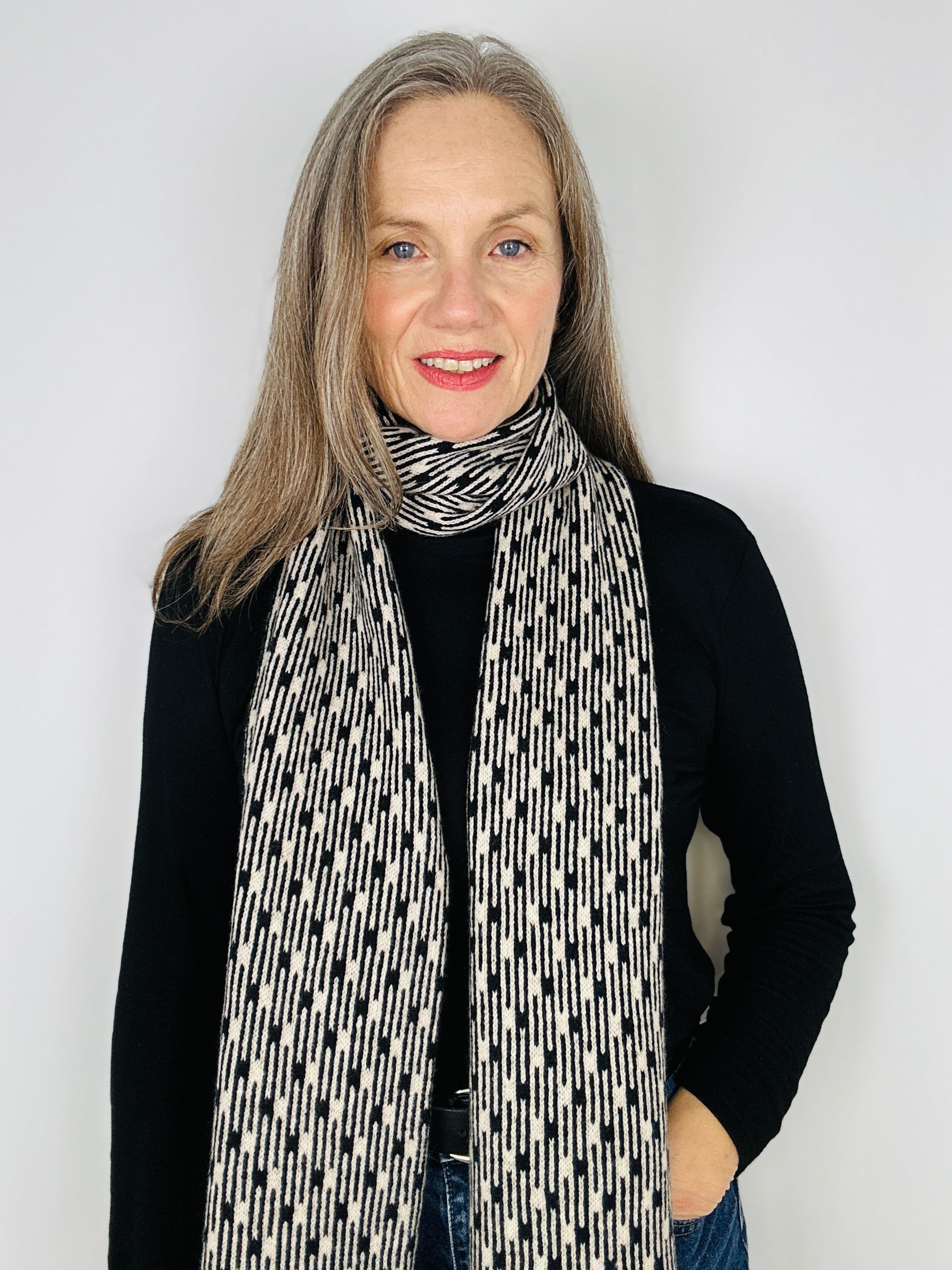 Beautiful sexy woman in a black top wearing a Black and Oatmeal Metropolis geometric patterned handcrafted merino lambswool scarf on a neutral backdrop.  Pom Shanty Knitwear.