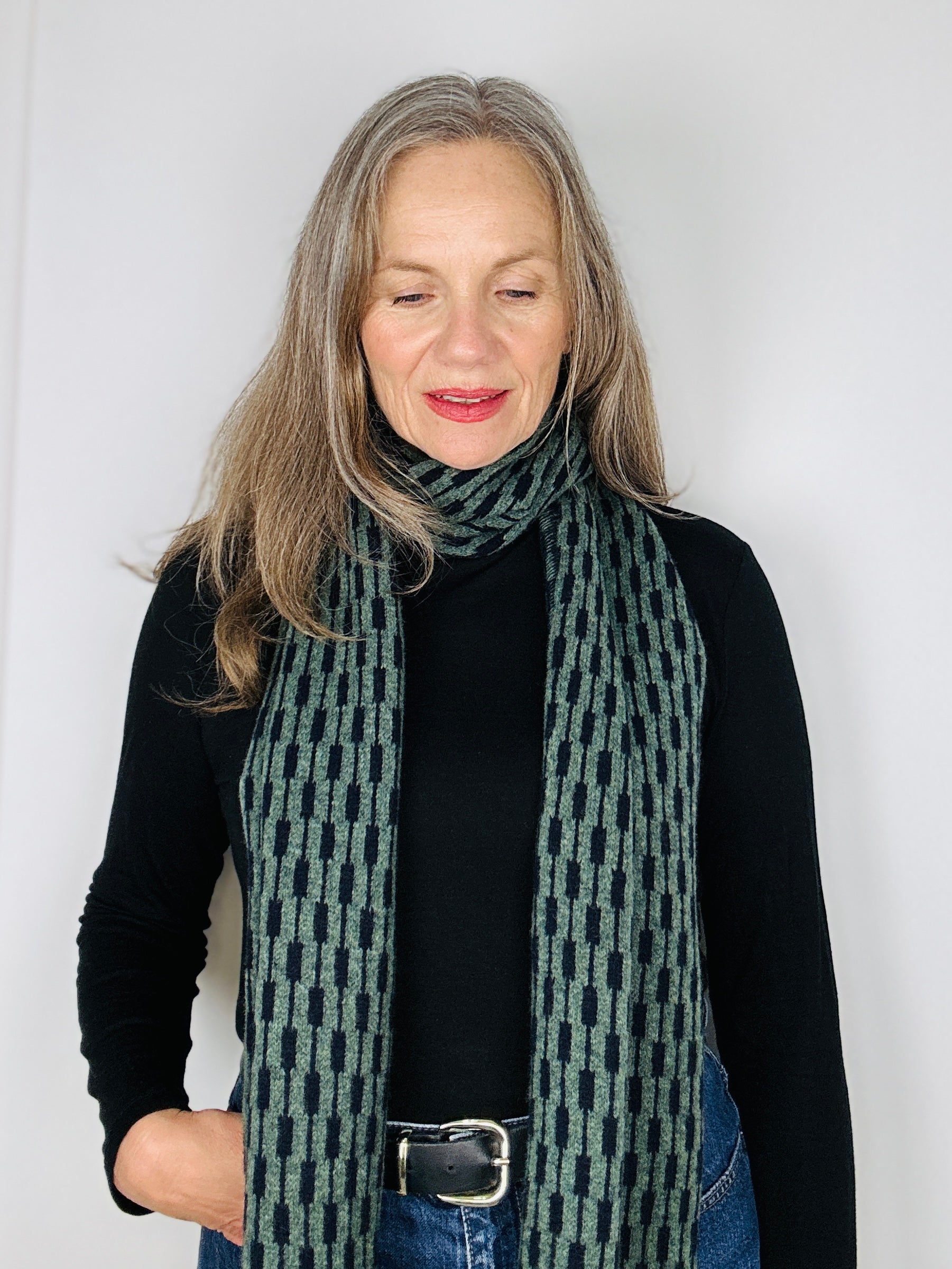 beautiful sexy woman in a black top wearing a peacock green and naby blue merino lambswool geometric patterned scarf on a plain background.