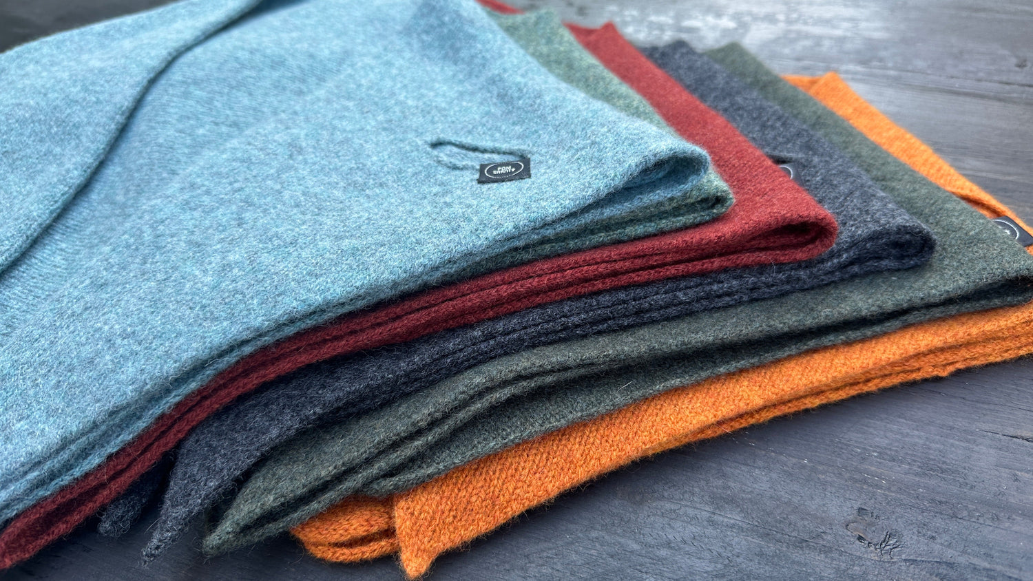 Stack of double-layer merino lambswool kerchiefs with pull-through slit in blue, green, sienna brown, charcoal, olive and rust.